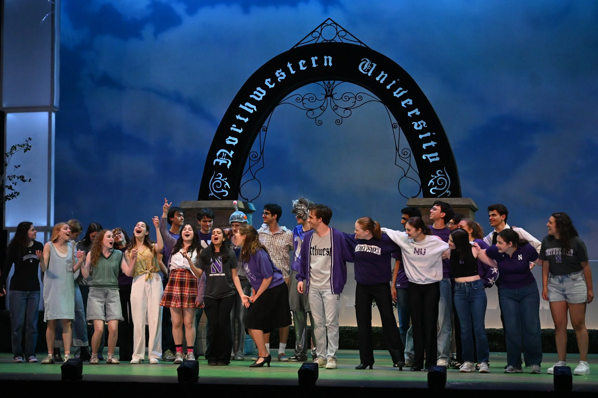 Group of students on stage at a university theater production, standing in front of an arch that reads 'Purdue Western University,' with a blue sky backdrop.