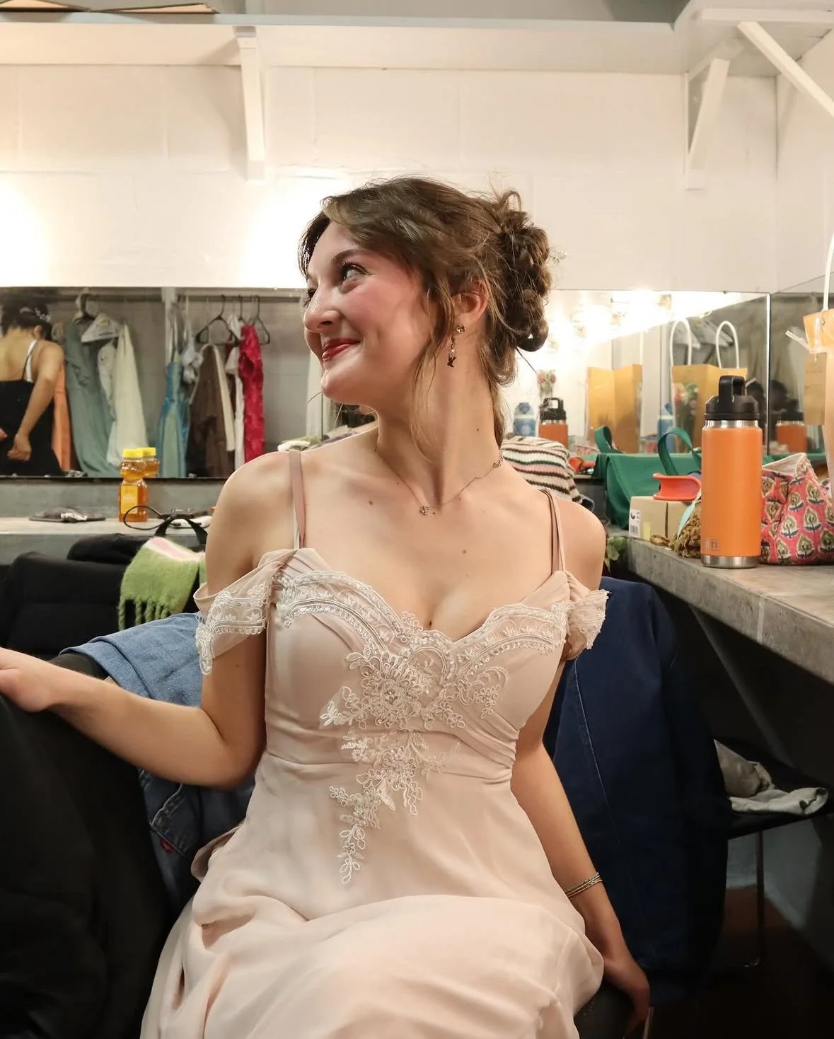 A woman sitting backstage in a dressing room with a cheerful smile, wearing a light pink dress with lace details, surrounded by clothing, bags, water bottles, and personal items.