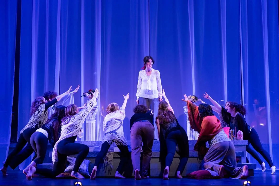 Stage scene with woman standing on platform, surrounded by women reaching towards her, set against blue curtains.