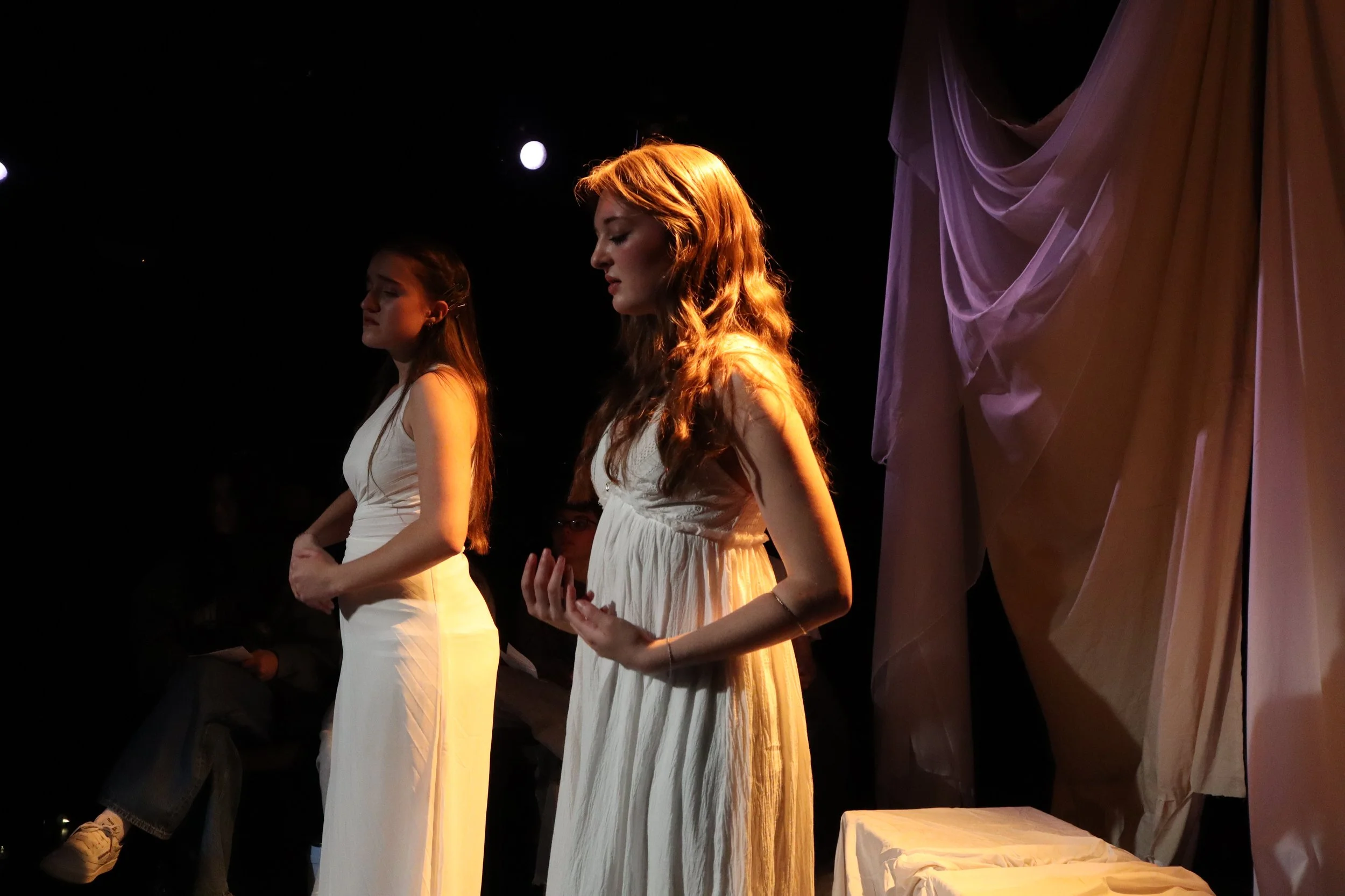 Two women in white dresses standing side by side on a dimly lit stage during a performance or rehearsal.