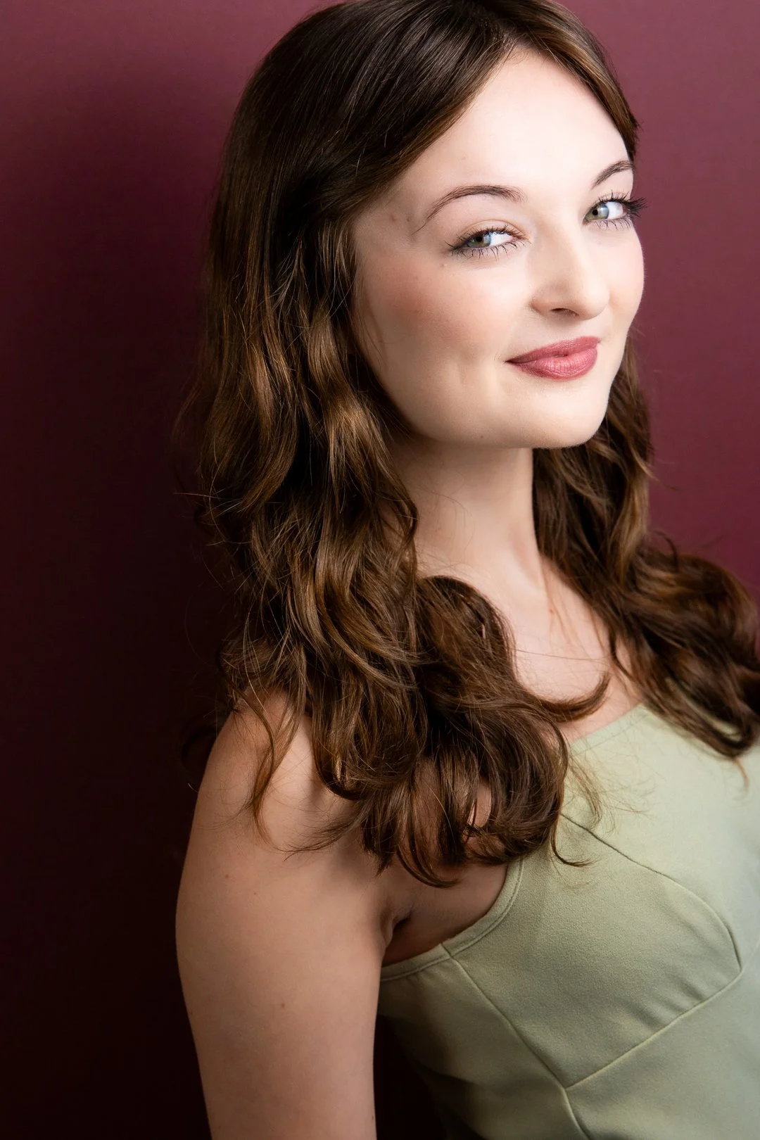 Headshot of Sarah Novak  with fair skin and long, wavy brown hair, smiling subtly, wearing a pale green top against a purple background.