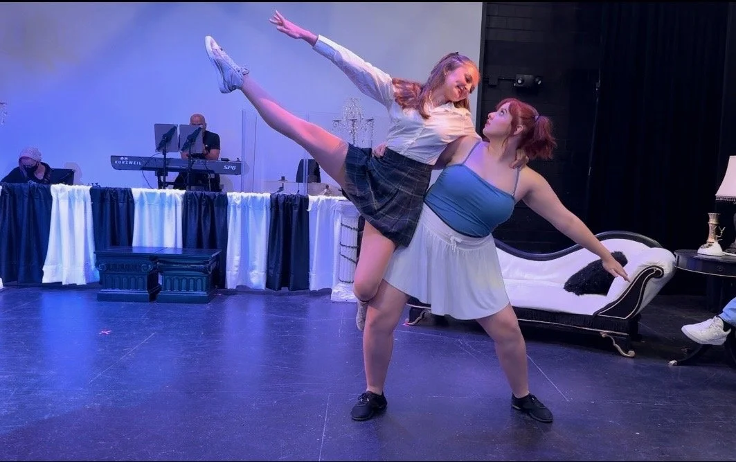 Two women performing a dance on stage, with one lifting the other. The background features music equipment and a decorative stage setup.