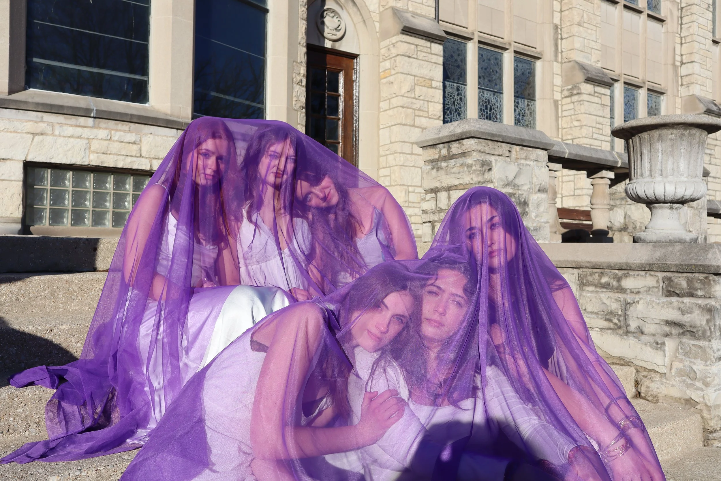 Five women sitting on outdoor stone steps, each covered with purple sheer fabric, in front of a stone building with large windows and decorative urns.