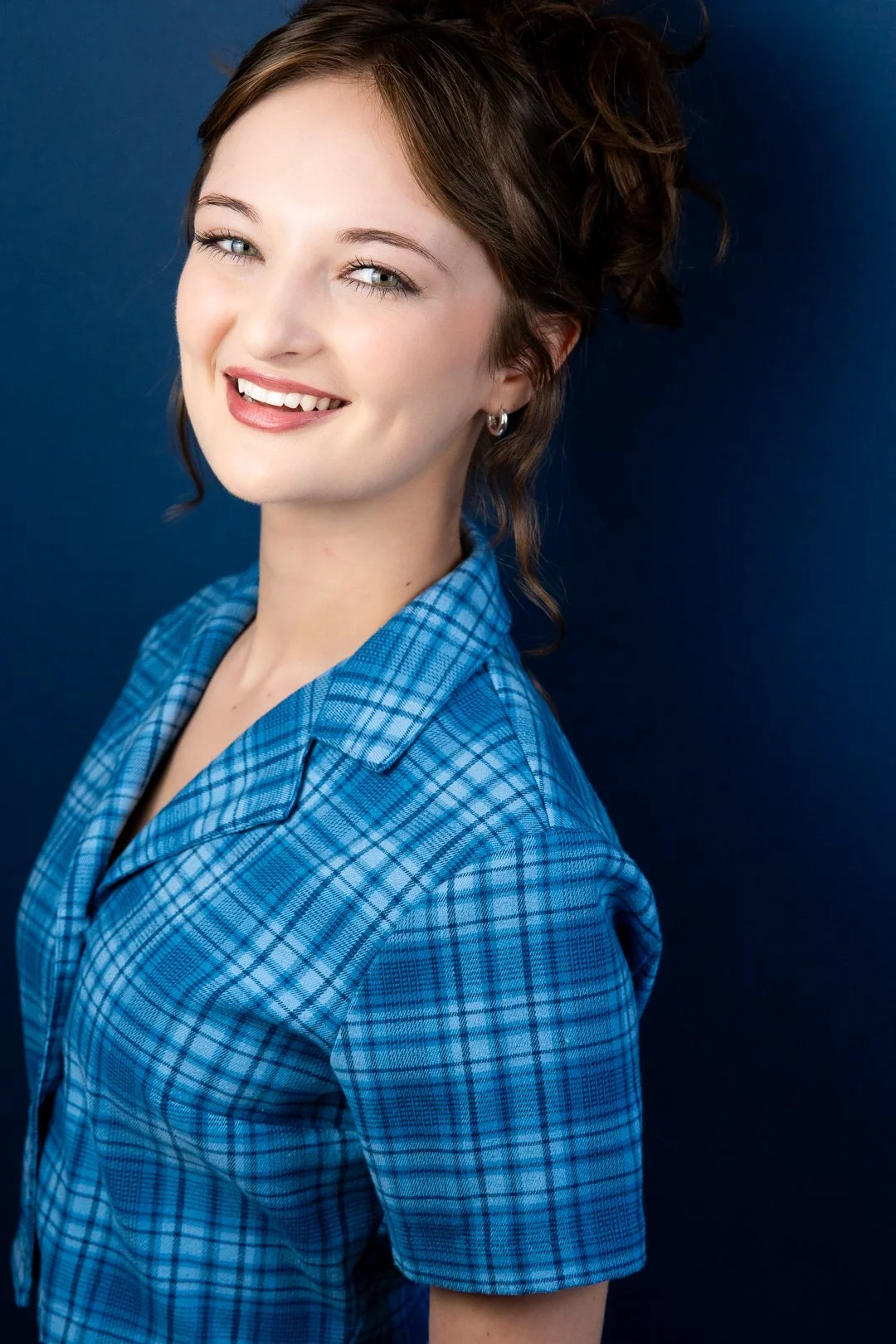 Headshot of Sarah Novak with light skin, brown hair styled in loose curls, wearing a blue plaid shirt, smiling, standing against a dark blue background.