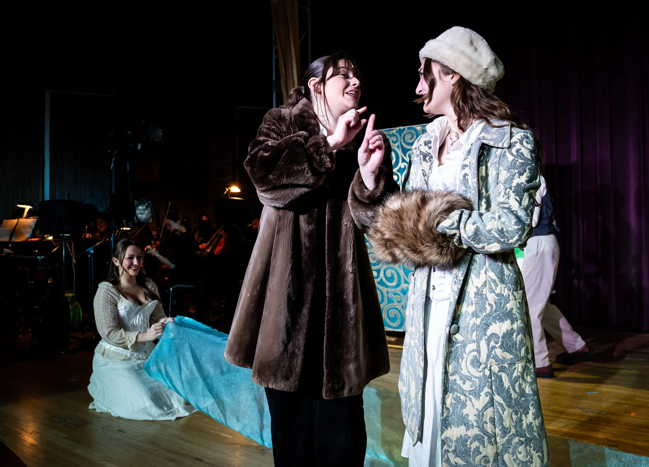 Three women on stage performing in a play, with two women standing close and one woman sitting on the floor, in front of a musical orchestra.