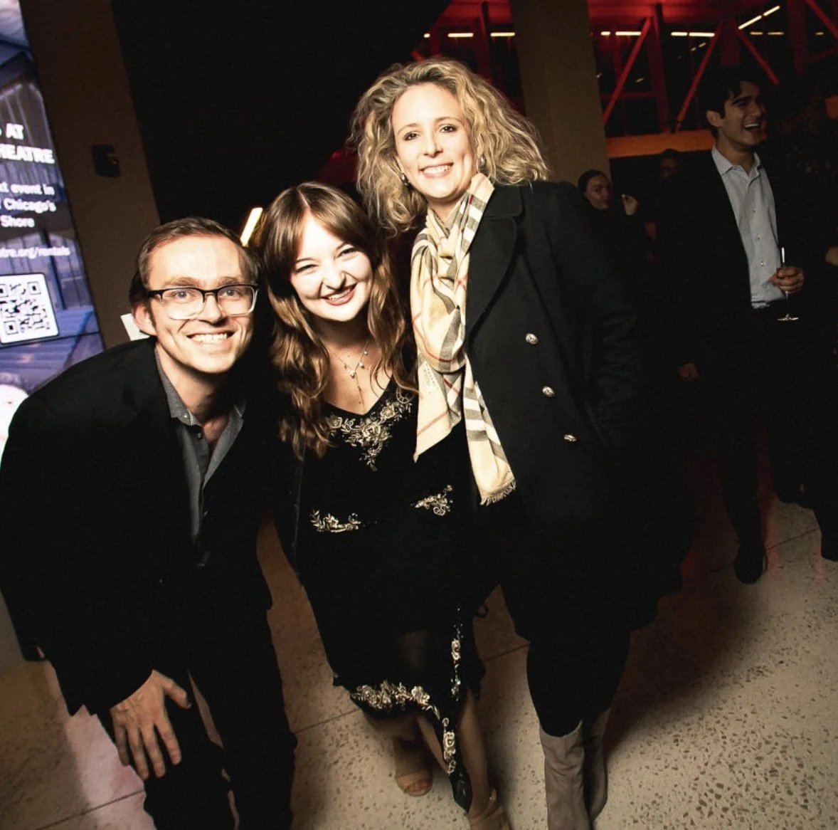 Sarah Novak, Mason Moss, and Heather Boehm at the As You Like It opening night party for Writers Theatre.Three smiling people, two women and one man, posing together at an event with a dark background and red lighting.