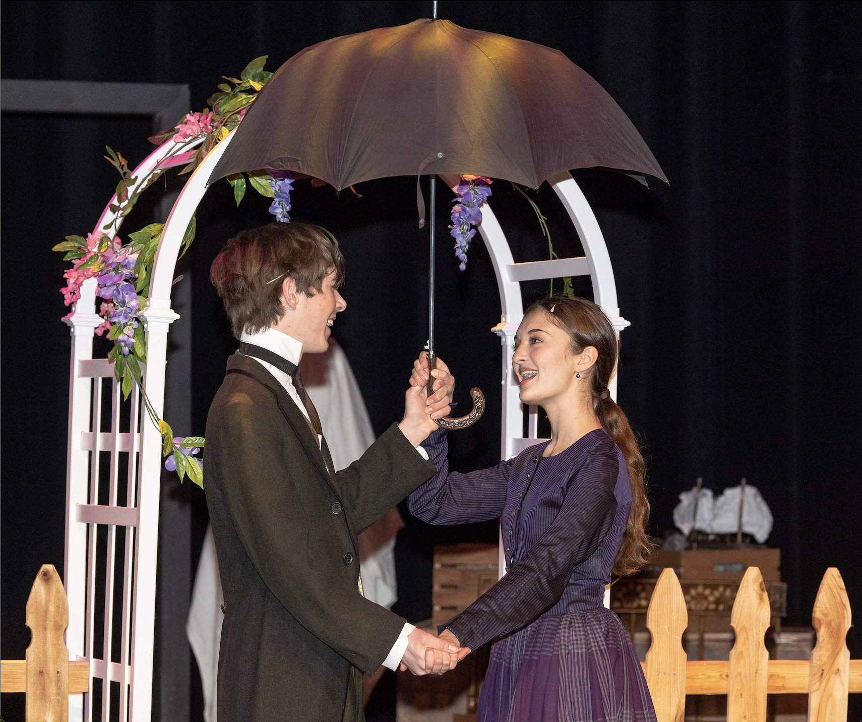 A young man and woman holding hands and smiling under a large black umbrella during a wedding or a formal event. They are standing under a decorated arch with purple and pink flowers, with black curtains in the background.