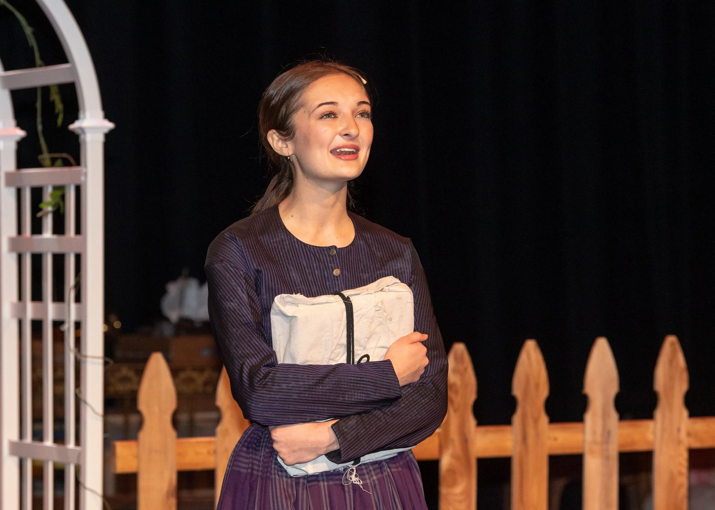 A young woman with brown hair tied back, holding a cloth bag and a book, standing outside near a wooden fence and white arbor, smiling and looking upward.