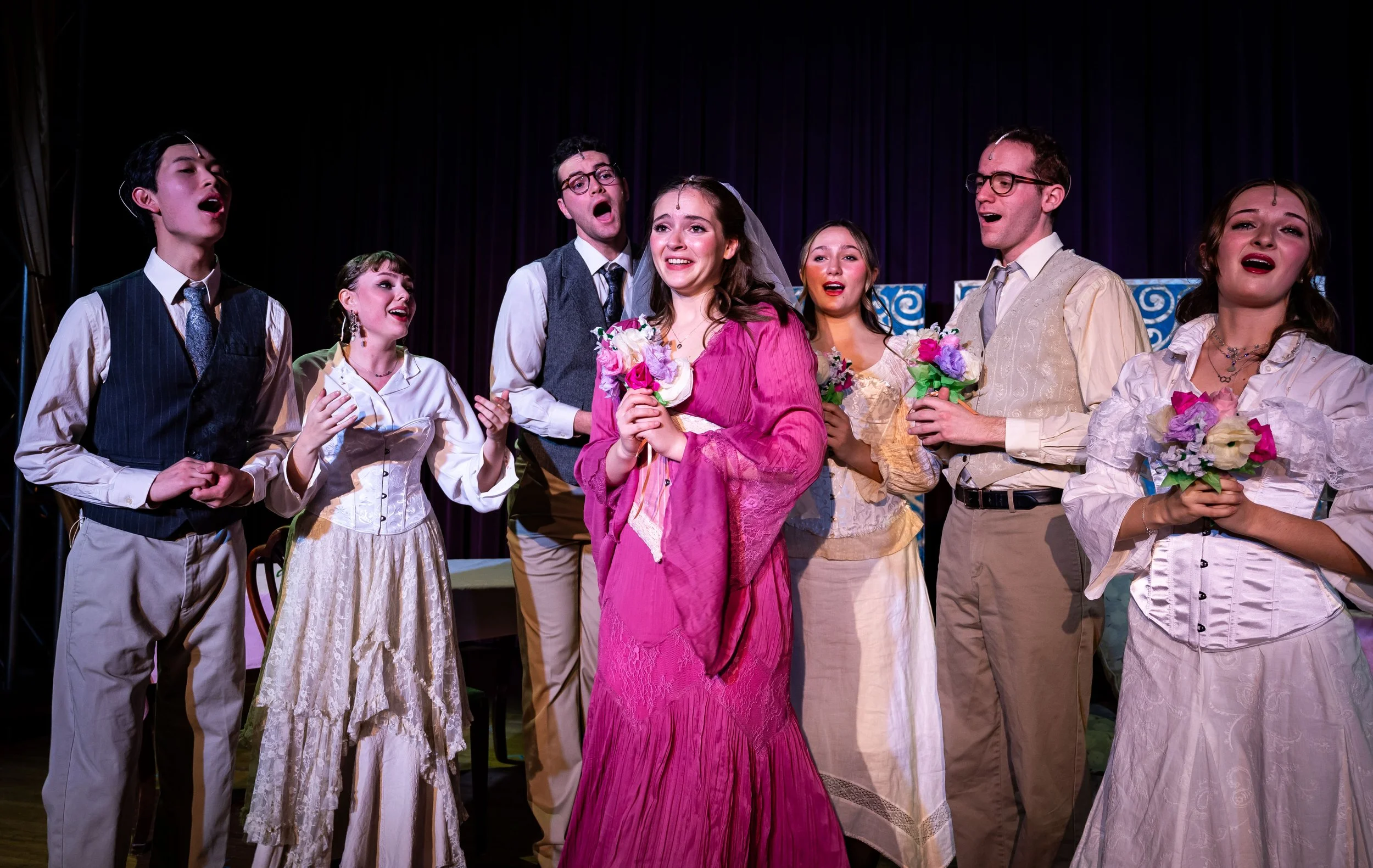 Actors performing on stage in a play, dressed in vintage clothing, holding bouquets of flowers, with a dark curtain in the background.