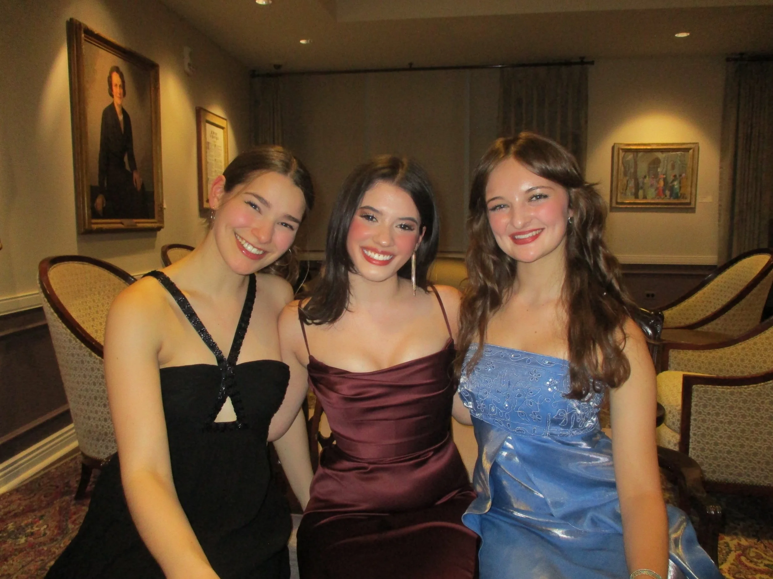 Three young women in evening gowns smiling and sitting close together in a room with artwork on the walls.