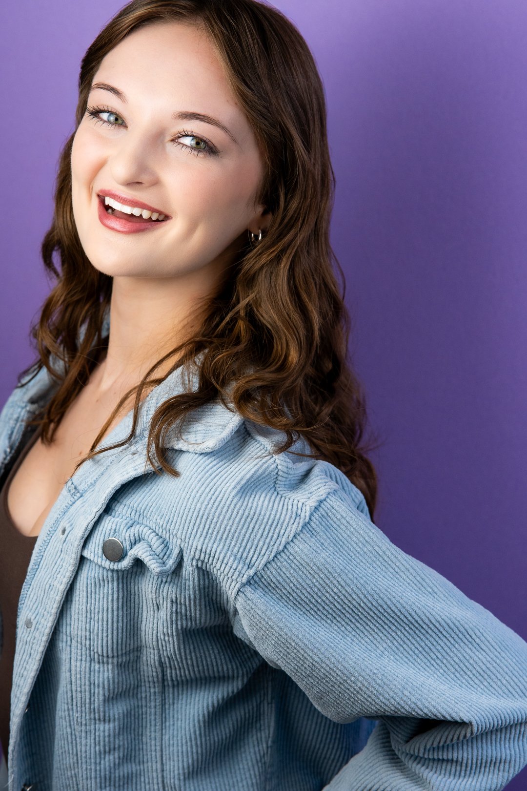 Headshot of Sarah Novak  with curly brown hair, blue eyes, and earrings, smiling at the camera against a purple background, wearing a light blue denim jacket over a black top.