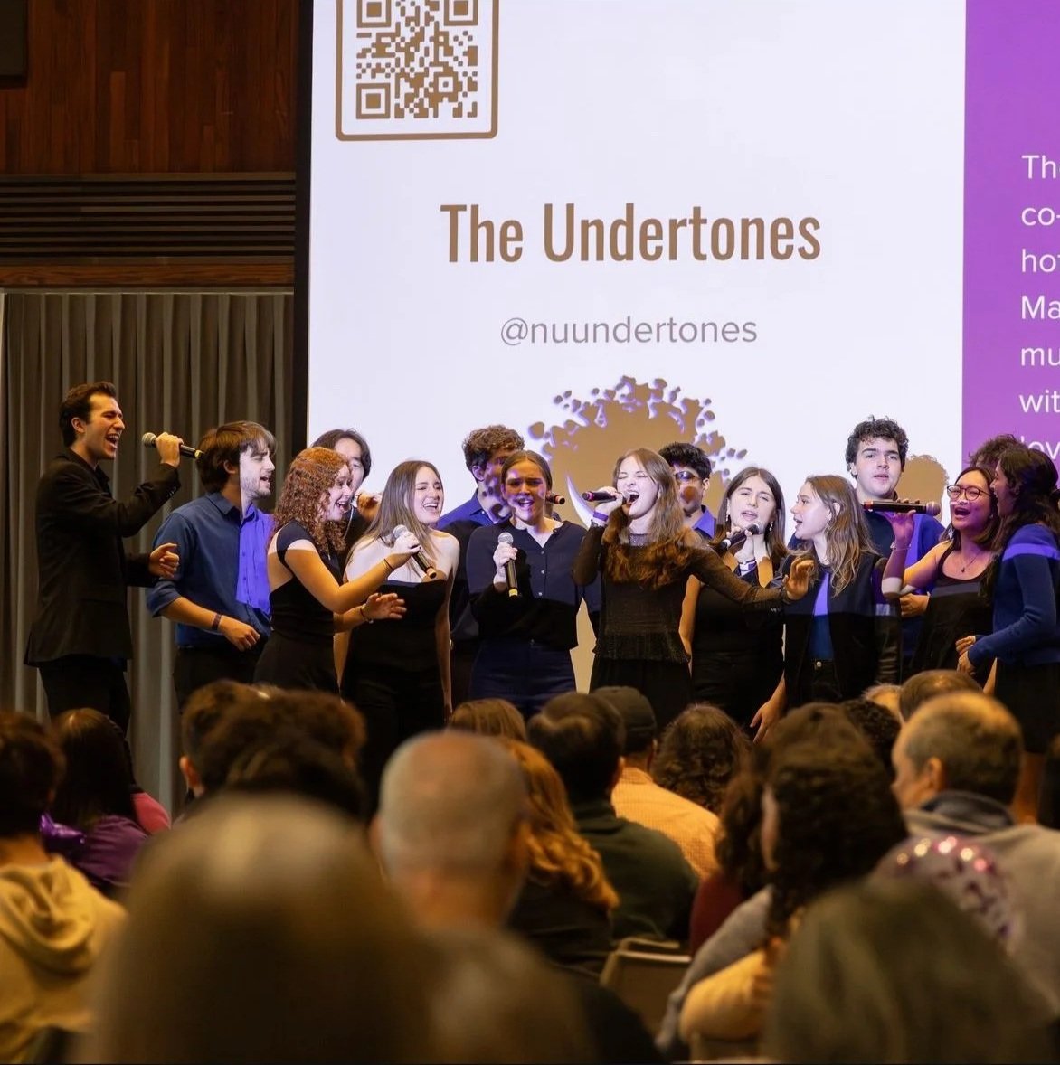 Sarah Novak performing with the Northwestern Undertones. Performers singing and dancing on stage at an event, with an audience watching. A large screen behind them displays the name 'The Undertones' and the Instagram handle '@nuundertones'.