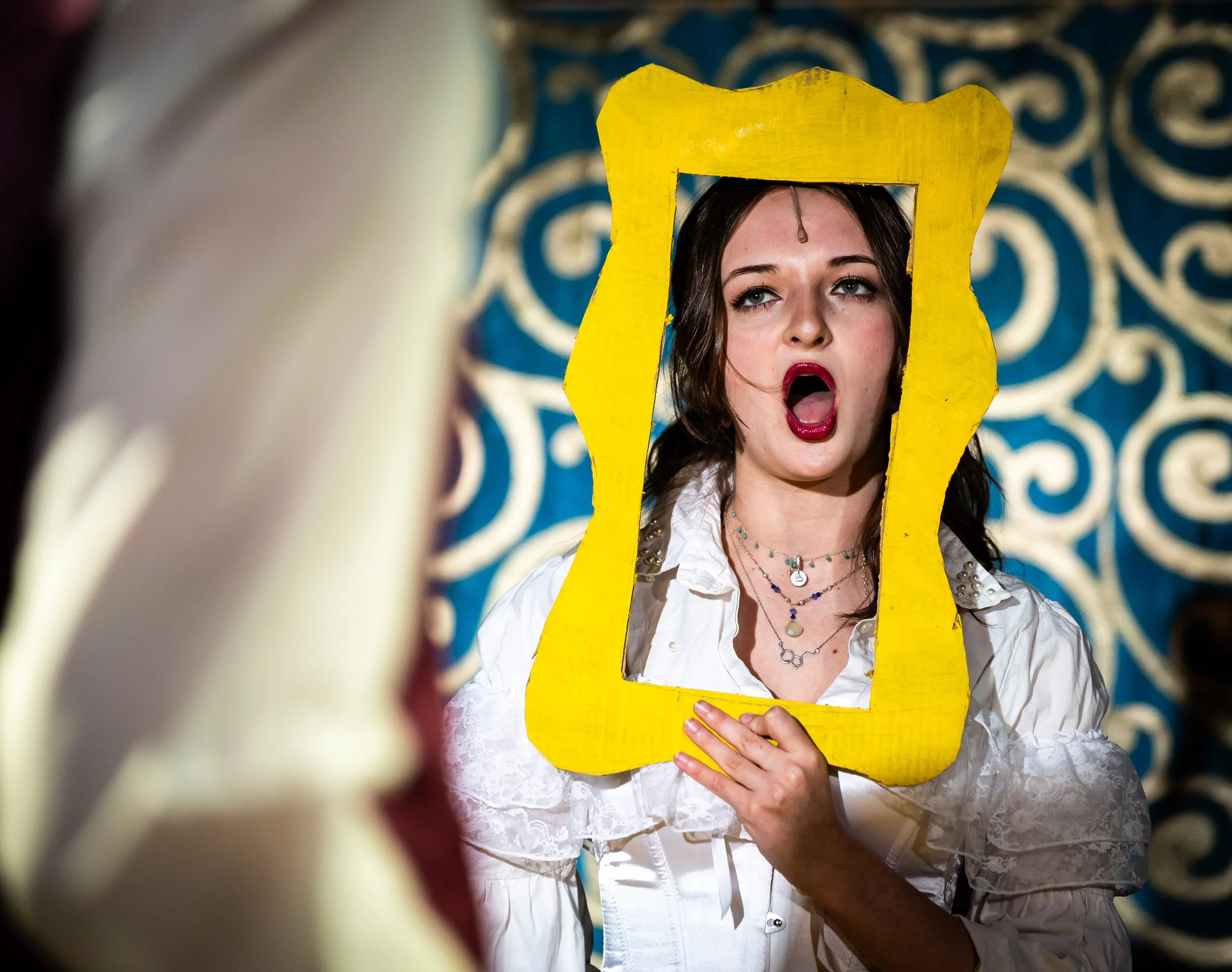 A woman looking into a yellow picture frame held in front of her face, with an animated facial expression of surprise or shock, in front of a decorative blue and gold patterned background.