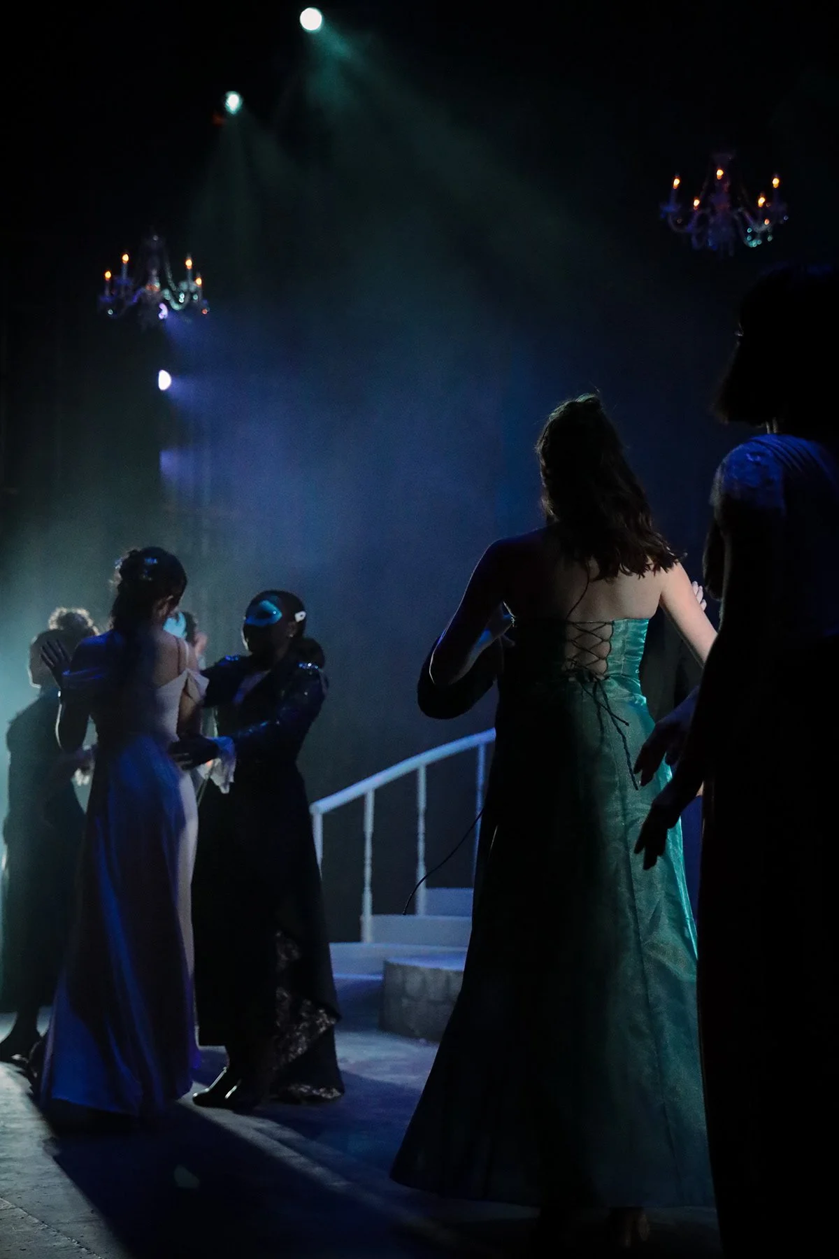 Silhouettes of women in long dresses on a dimly lit stage with chandeliers overhead.