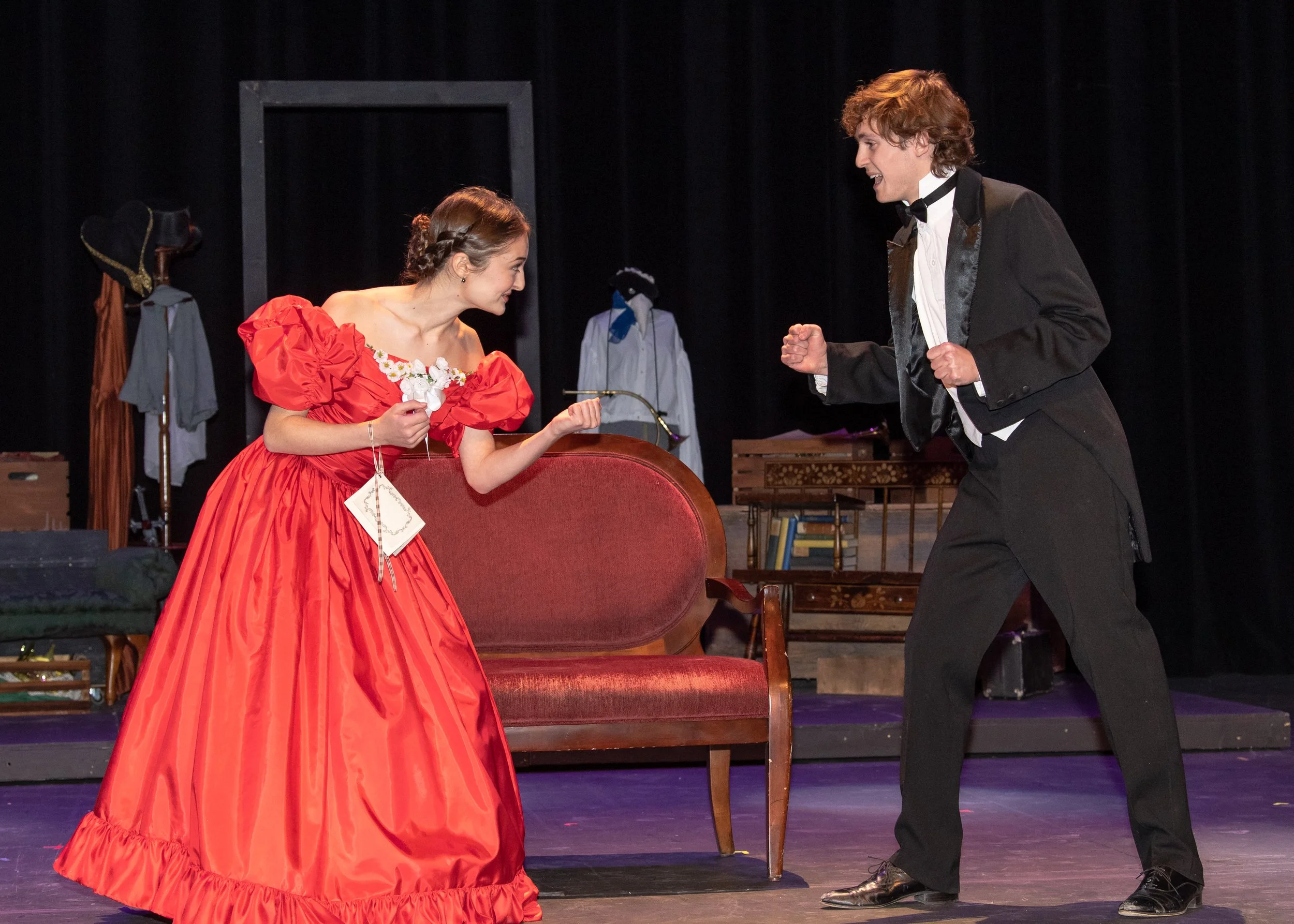 Two actors performing on stage in a play. The woman in a red dress with puffed sleeves is smiling and holding a flower, while the man in a tuxedo is gesturing energetically towards her. The stage has a vintage furniture setting with hats and clothes in the background.