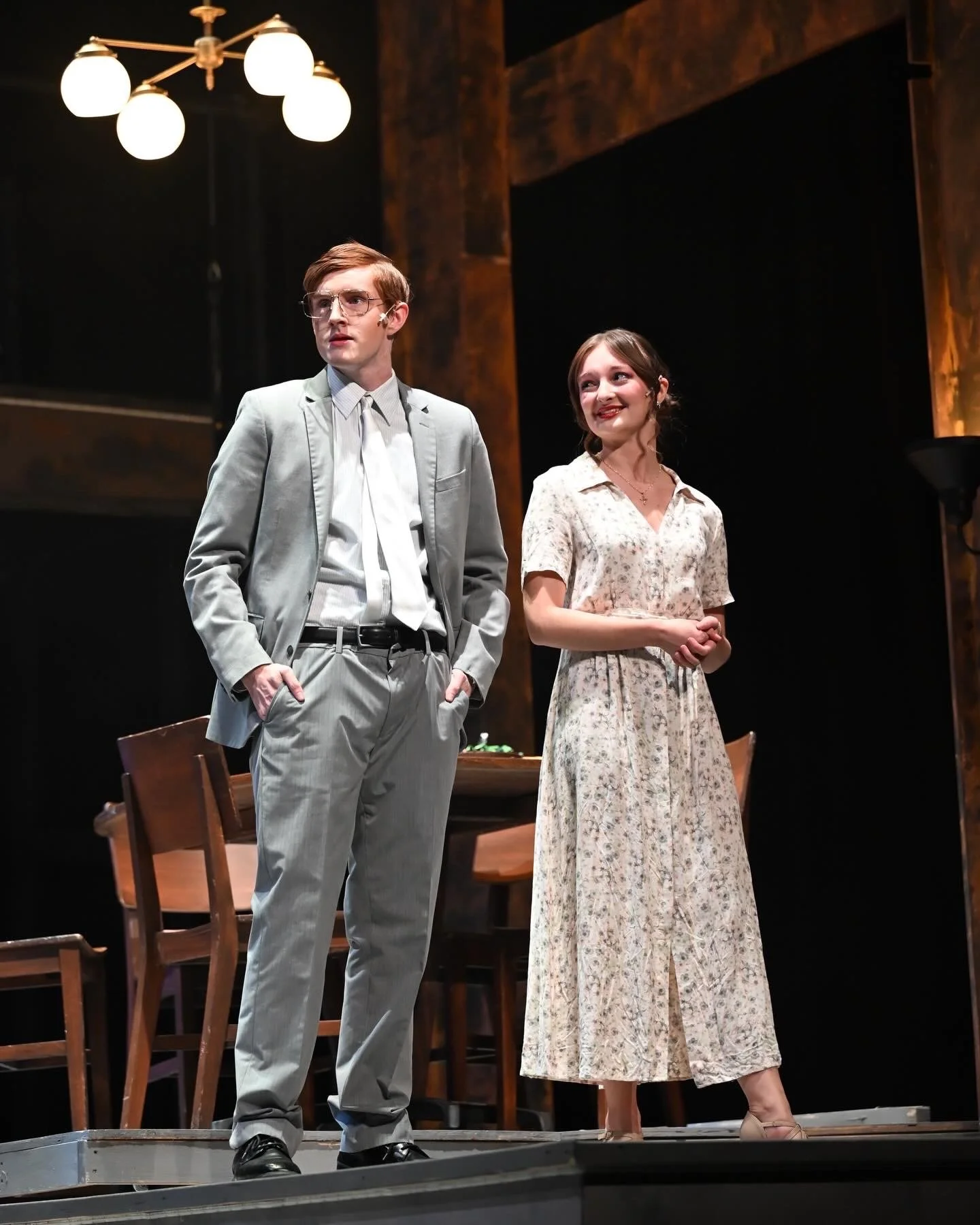 Sarah Novak and Garraway Nance on stage during a theatrical performance. The man is wearing glasses, a light gray suit, and a white shirt, with hands in his pockets. The woman is wearing a floral dress and smiling.
