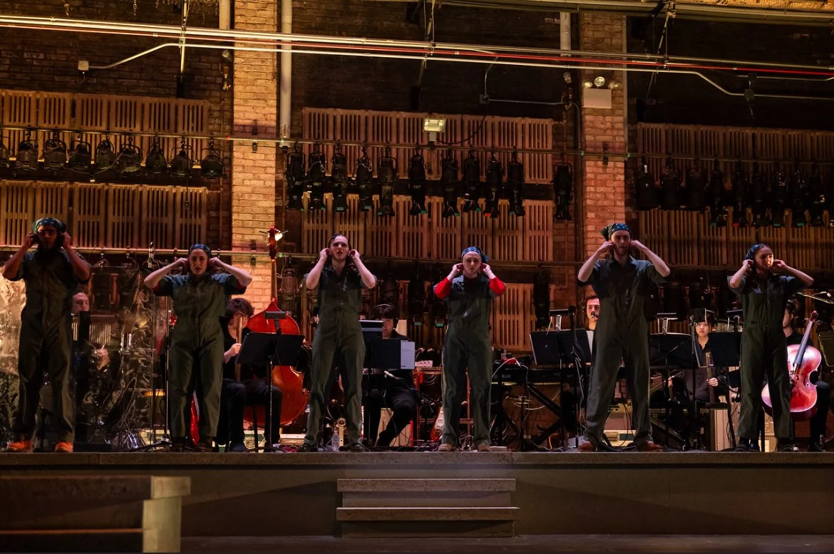 A group of six performers standing on stage, wearing green coveralls and headphones, with a small orchestra and musical instruments behind them in a recording studio or theater setting.