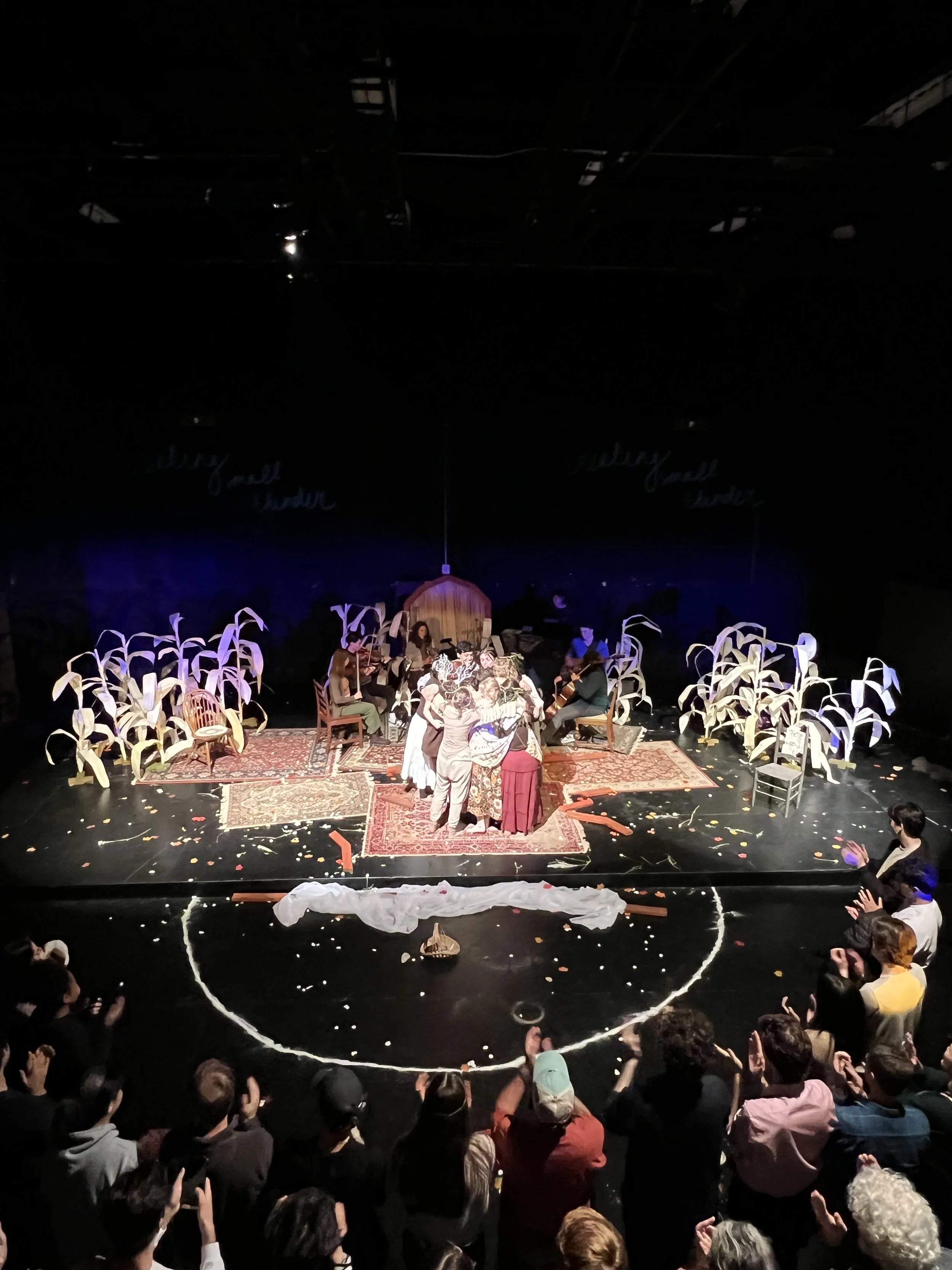 Theater stage with performers and an audience clapping, decorated with corn stalks, rugs, and props for a play or performance.