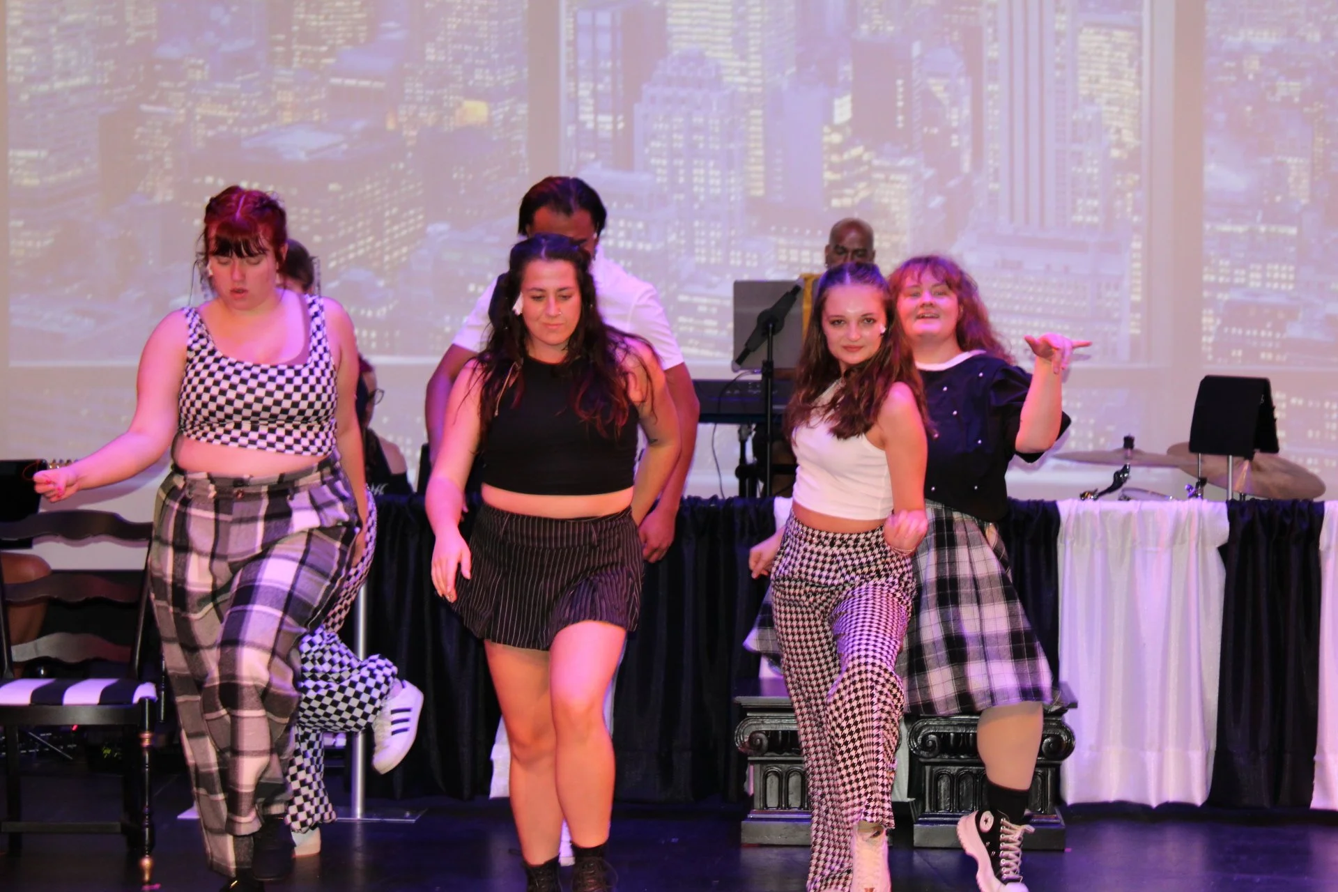 Four young women dancing on stage with a DJ in the background and a city skyline projected behind them.