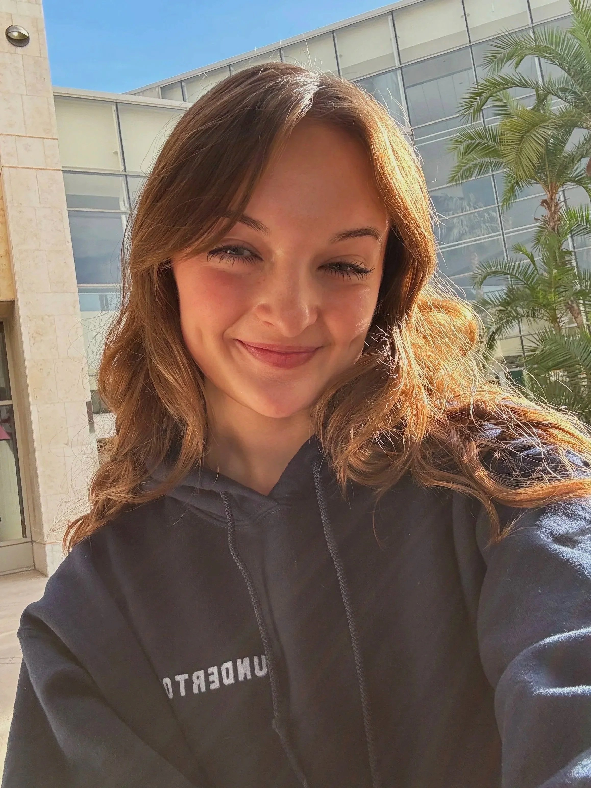 Sarah Novak with light brown hair smiling while taking a selfie outdoors. She is wearing a dark hoodie, standing in front of a modern building with glass windows and palm trees.