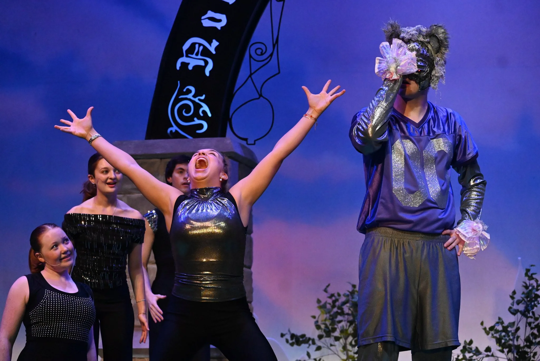 A theatrical stage scene with five performers. One woman in a shiny black or dark gray dress with arms raised and mouth open, appears excited or triumphant. Other women in black costumes stand nearby, looking at her. A man in a colorful costume with a mask and elaborate headpiece, decorated with a large pink bow, stands to the right. A backdrop features a dark decorative structure with symbols or writing.