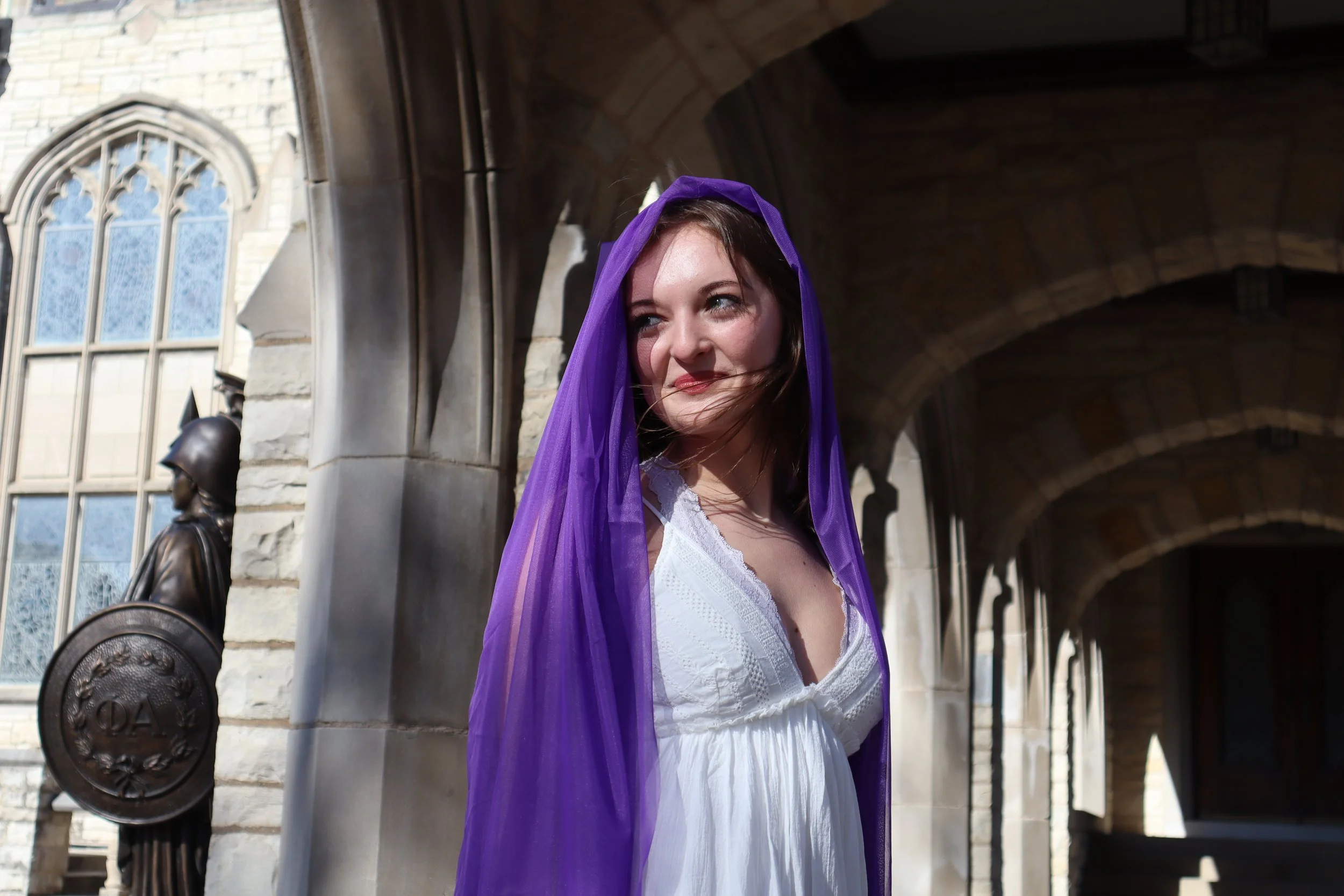A young woman in a white dress with a purple sheer shawl draped over her head, standing in front of a stone building with arches and a bronze statue of a soldier.