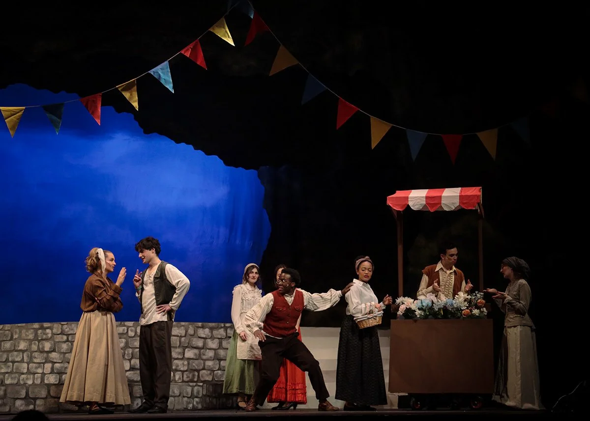 Theatrical stage scene with seven actors in period costumes, colorful bunting overhead, a blue screen backdrop, and a flower stand with a vendor, depicting a lively play.