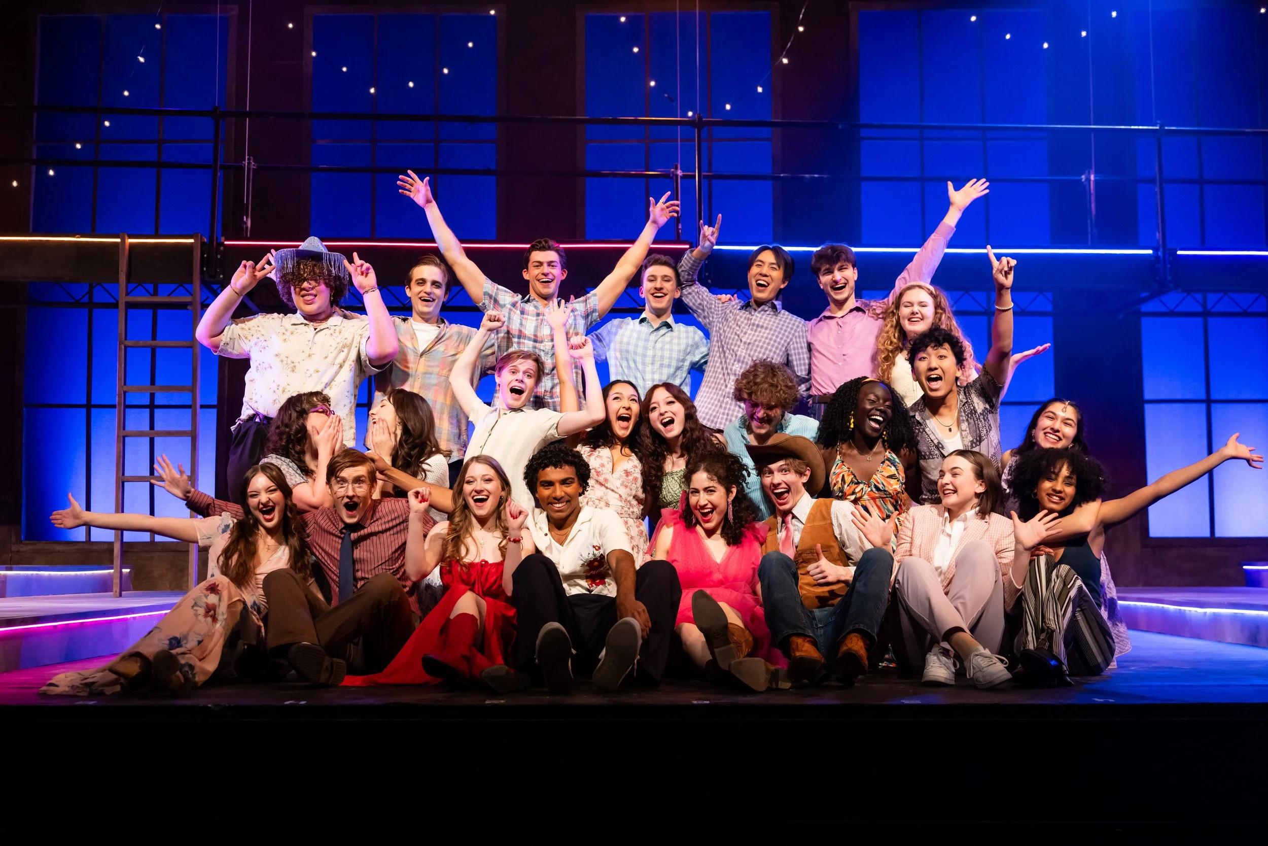 The cast of Footloose at Northwestern University on stage celebrating after a performance, with some sitting and others standing, in a theater with colorful lighting.