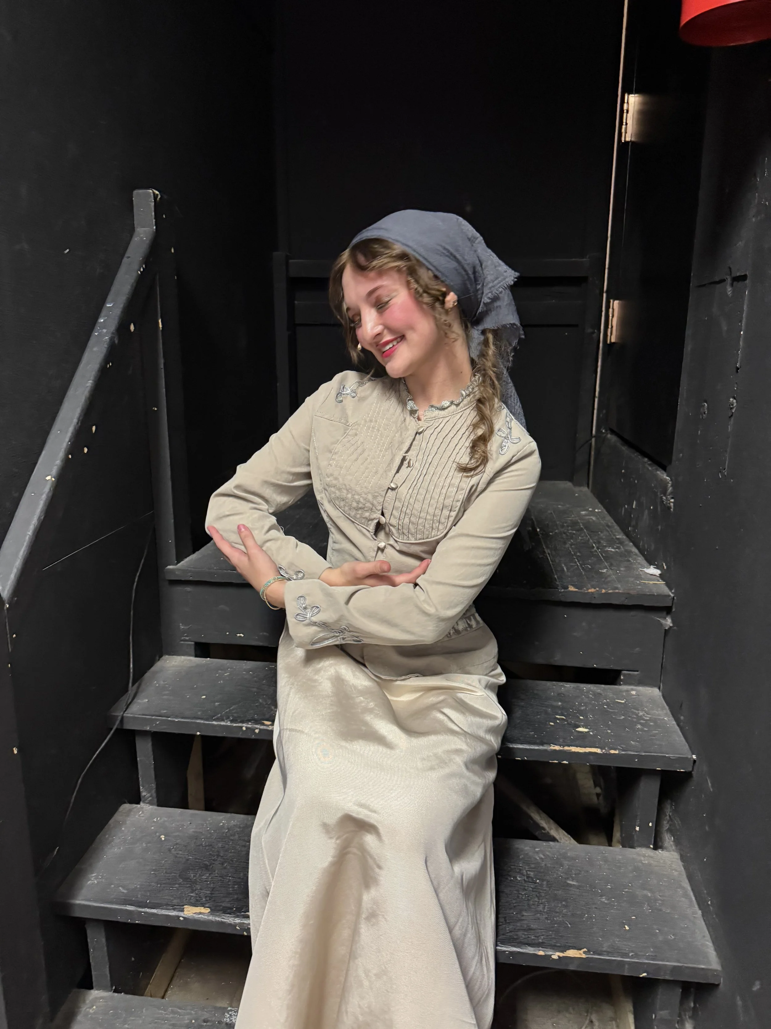 Sarah Novak in Cinderella at Northwestern University. Smiling woman wearing a beige embroidered dress and gray headscarf sitting on black wooden stairs in a dark room.