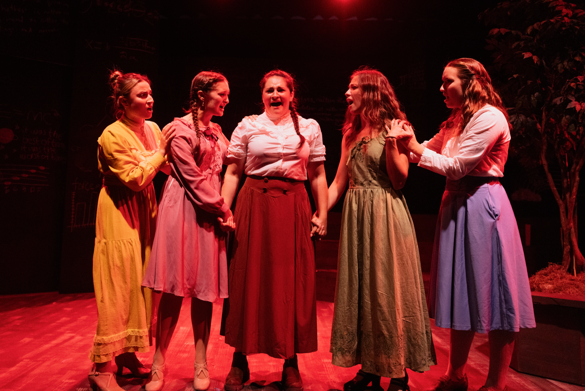 Six women on stage in period costumes holding hands, appearing emotional and distressed, with a red lighting background.