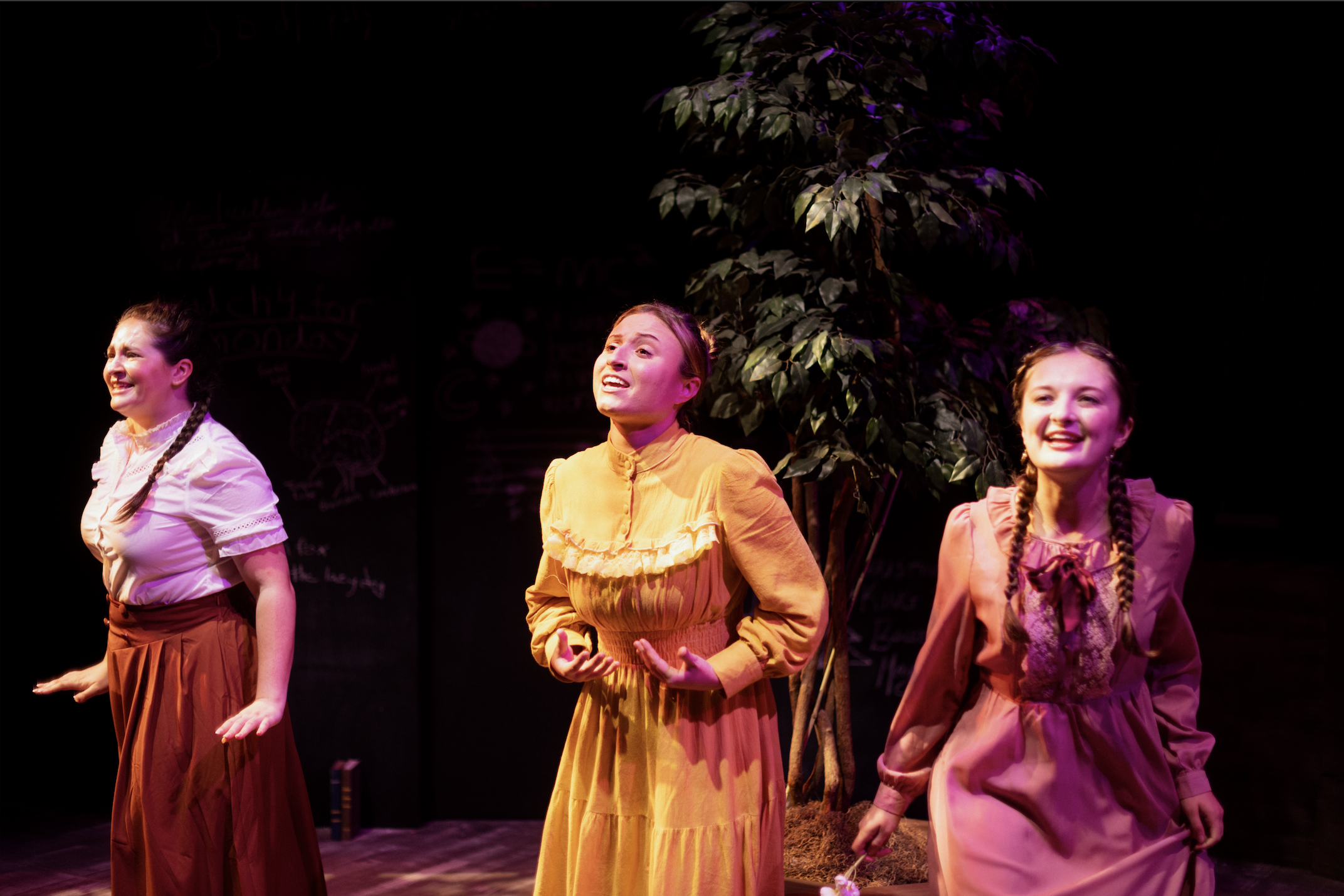 Three young girls performing on stage, dressed in period costumes, with a writing blackboard and a large plant in the background.