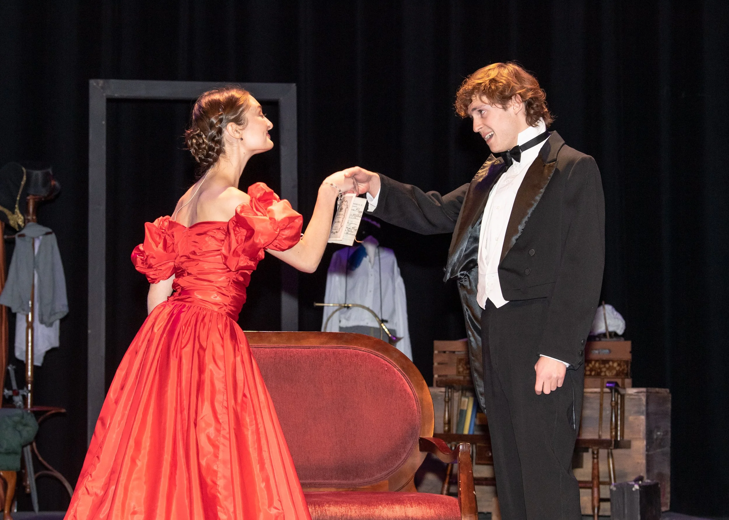 Two performers on stage, a woman in a red gown and a man in a tuxedo, sharing a joyful moment during a theatrical performance.