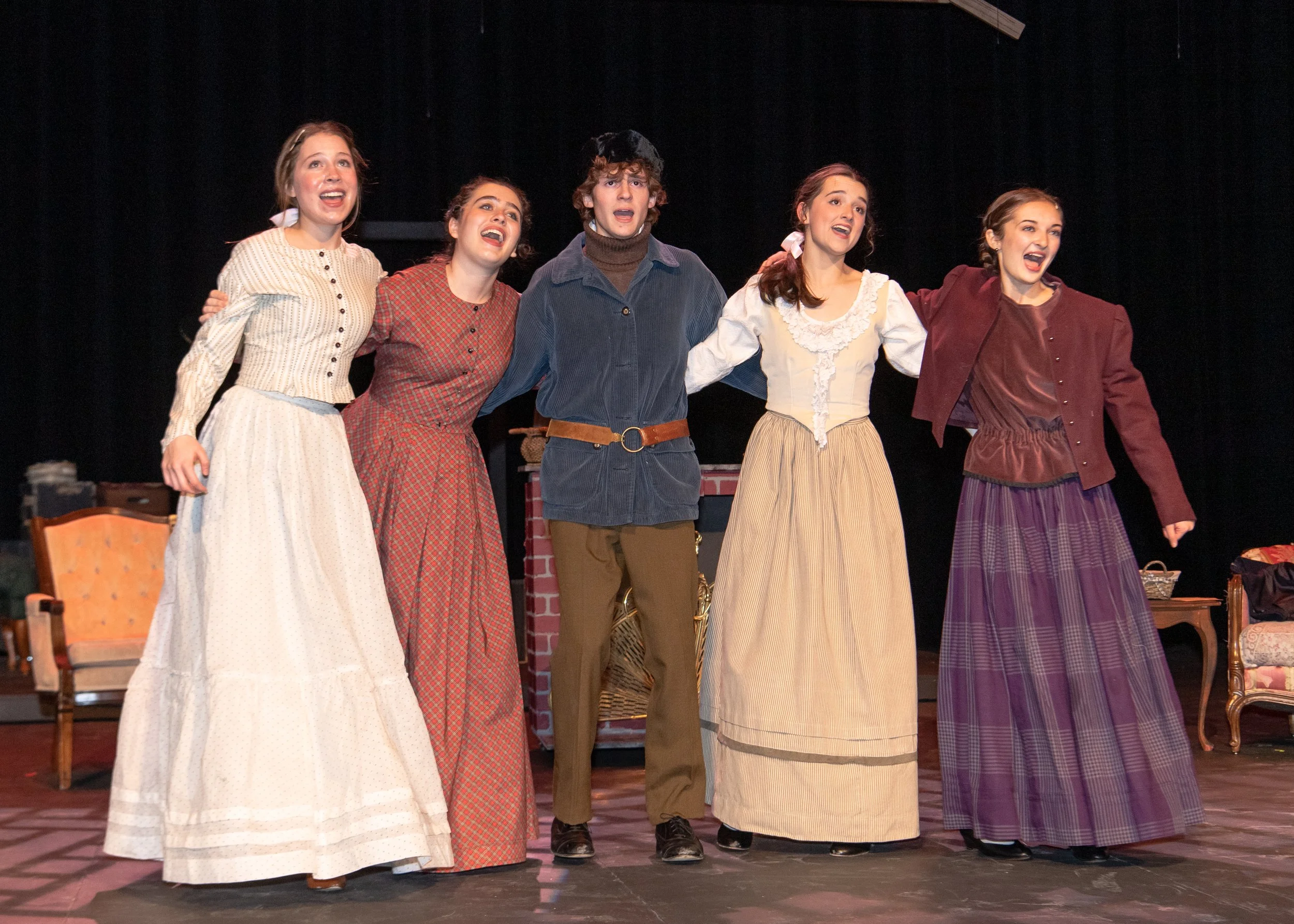 Five performers in period costumes on stage, taking a bow with arms around each other, after a theatrical performance.