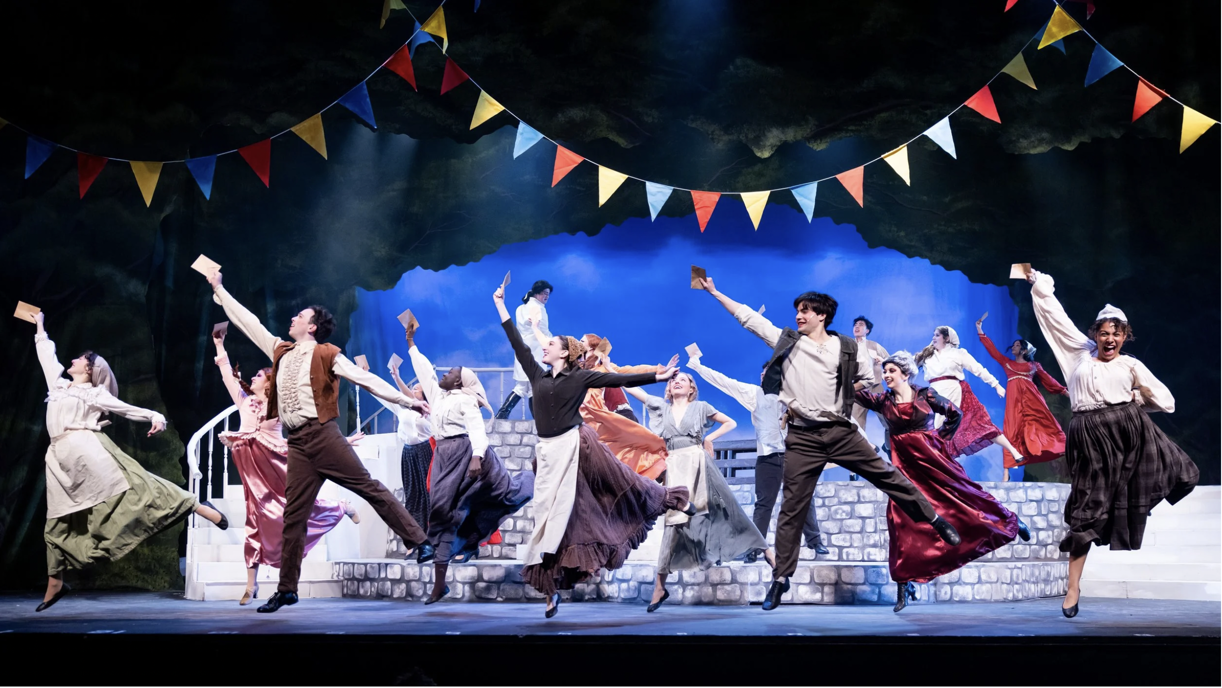 A group of performers dressed in period costumes joyfully dance on stage during a theatrical play, with colorful banners hanging above them and a set resembling a village square in the background.