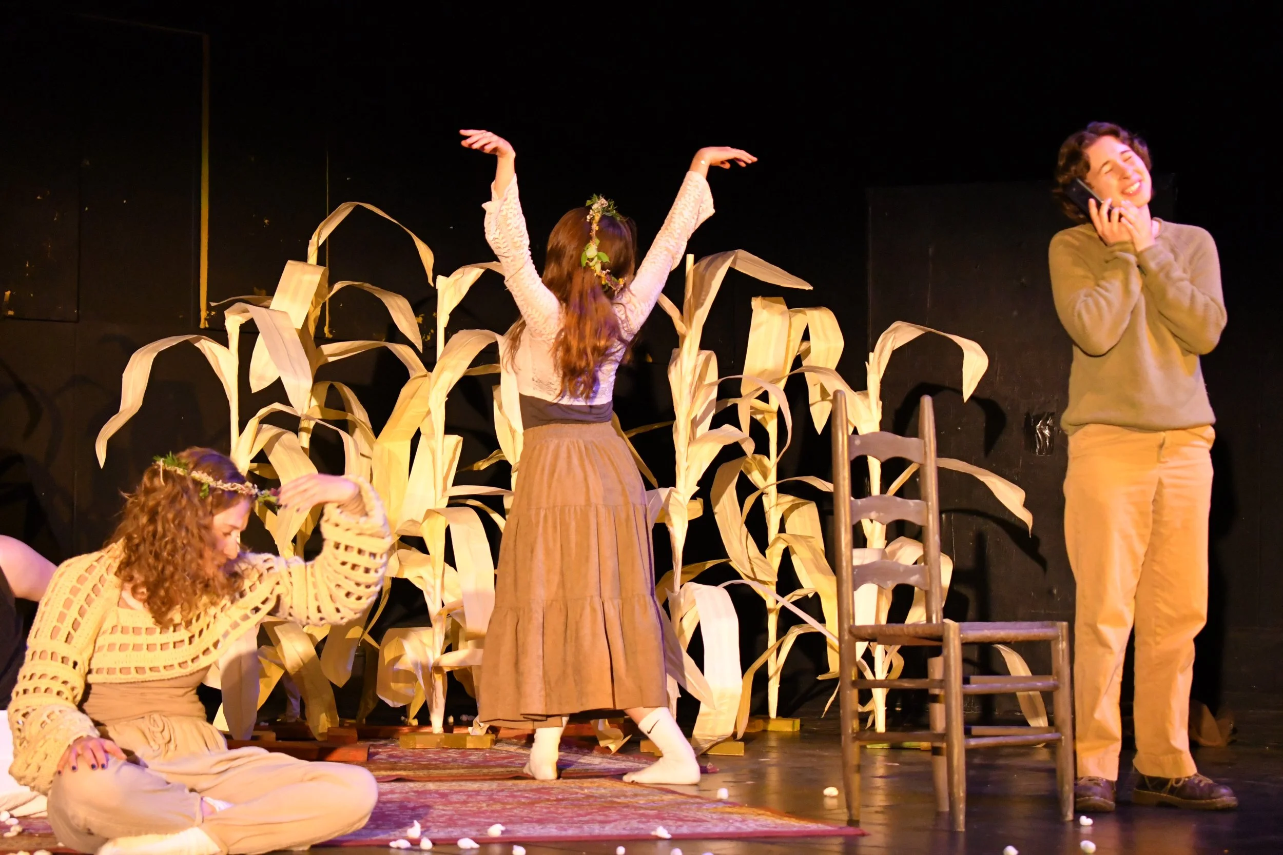 The image shows three women in a theatrical stage setting. One woman is sitting on the floor adjusting her hair, wearing beige pants and a crocheted sweater. Another woman is standing with her back to the camera, wearing a long skirt and a flower crown, raising her arms. The third woman, standing to the right, is on the phone, smiling, and wearing beige pants and a sweater. The background has a black wall with tall artificial corn stalks, and a wooden chair is situated on the stage.