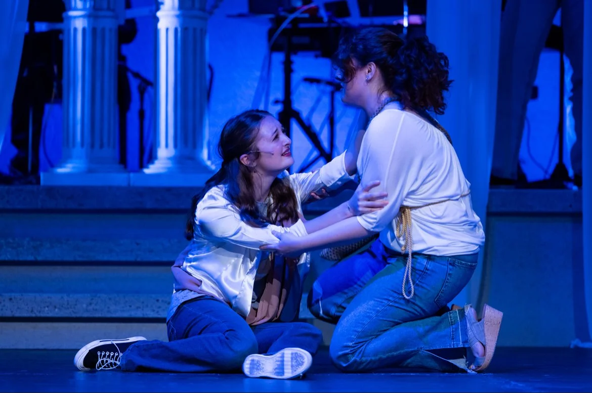 Sarah Novak in the Waa Mu Show at Northwestern University. One woman is sitting on the floor, looking up at the other woman who is kneeling and holding her by the shoulders. The background features stage props and blue lighting.