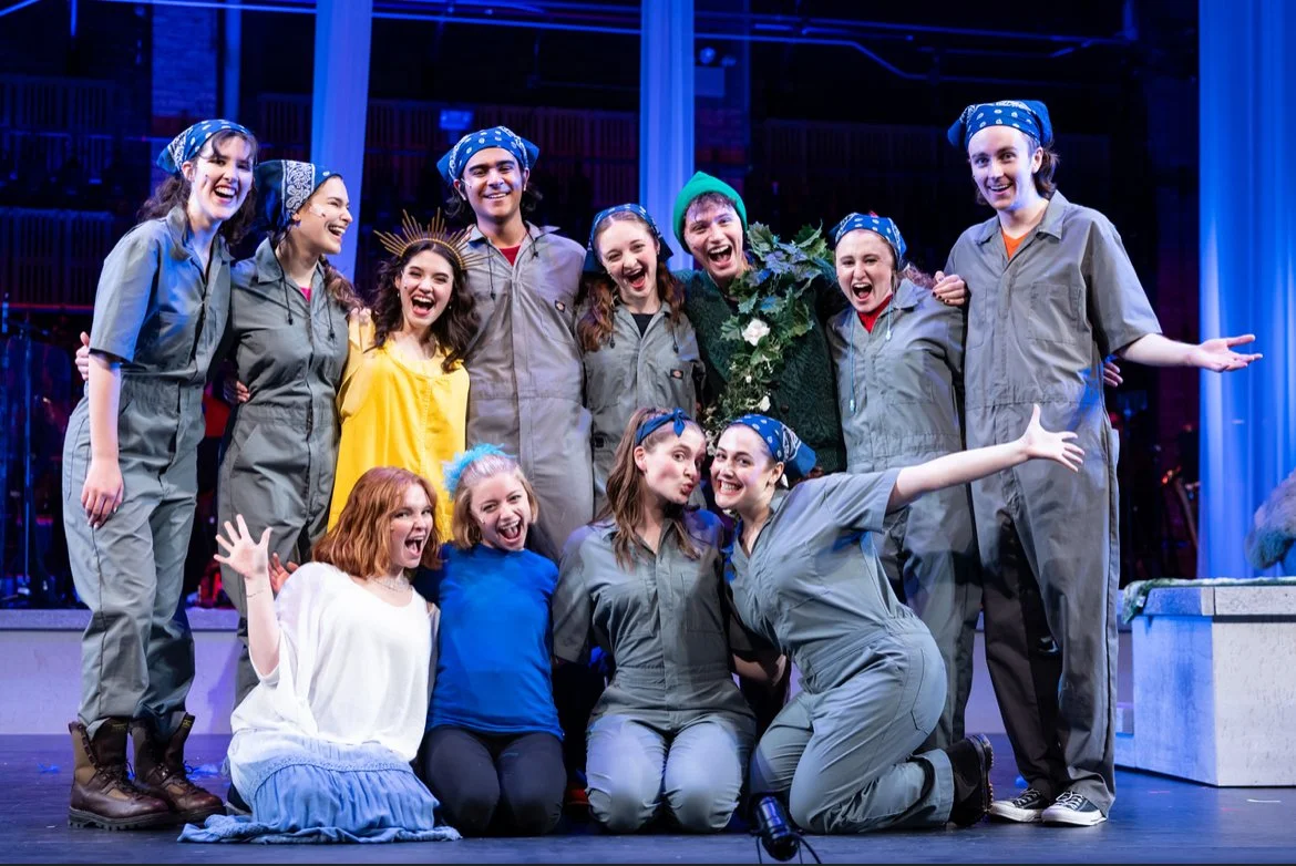 Cast members of a theatrical production celebrate on stage, dressed in costumes with many in gray jumpsuits and blue bandanas, some with headbands, standing together with arms around each other and smiling.