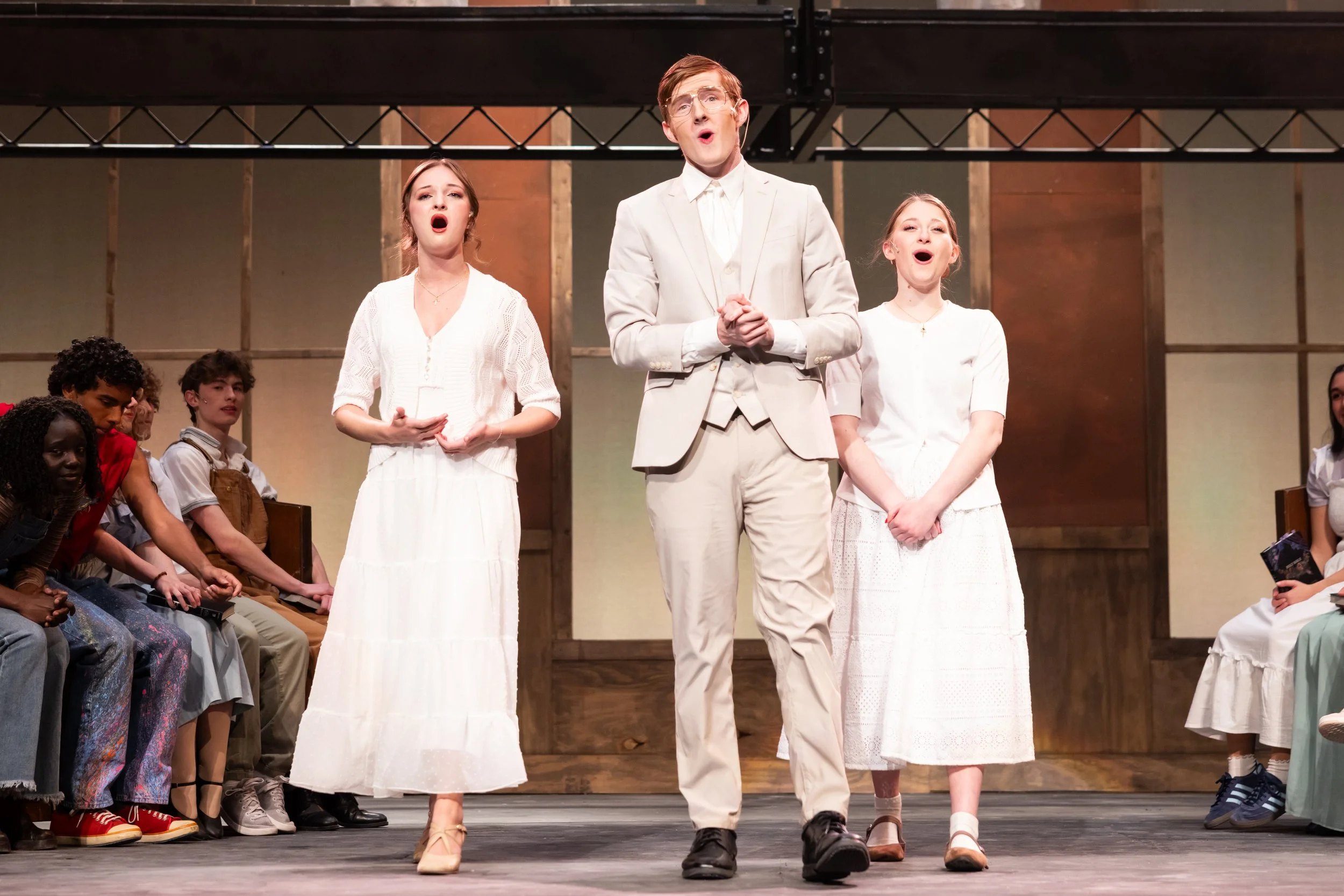 Sarah Novak, Garraway Nance, and Anna Rigo on stage singing, dressed in vintage white attire, with a group of diverse children sitting on benches watching, set against a wooden background.