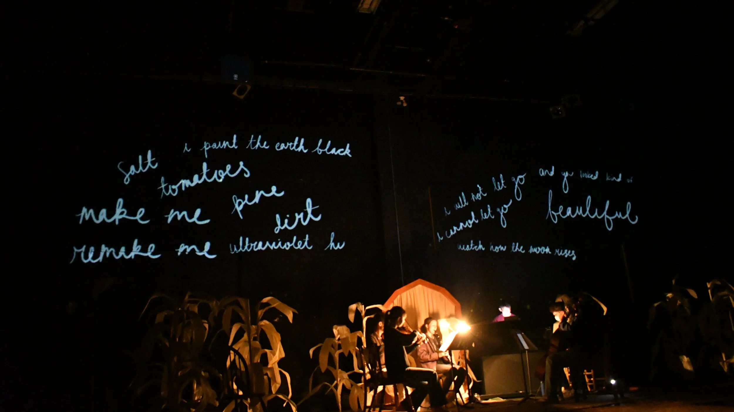 A theatrical stage features a group of performers sitting with musical instruments and music sheets, lit by warm stage lights. The background has black walls with handwritten lyrics projected in blue cursive text.