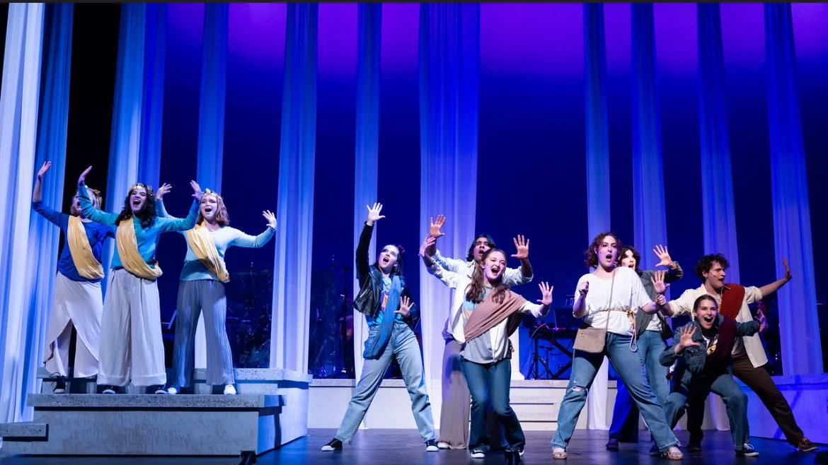 Group of young performers on stage during a theatrical production, with some singing and gesturing, colorful background lighting.