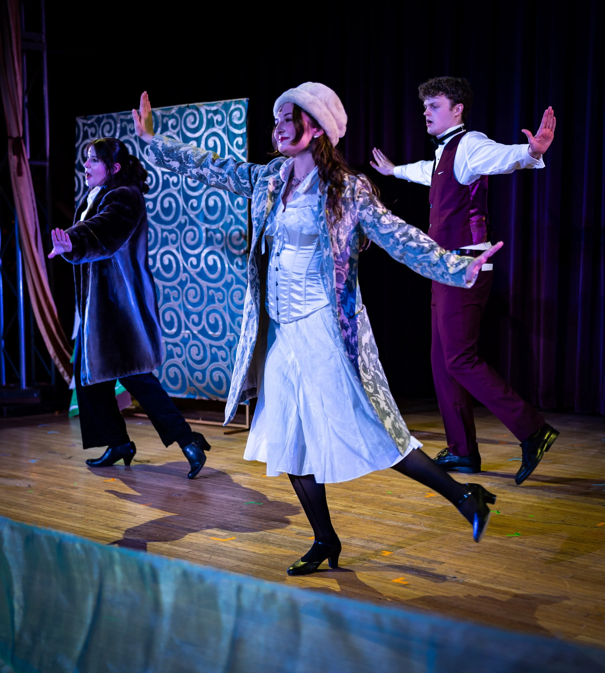 Three performers dancing on a stage with a colorful backdrop, wearing vintage costumes.