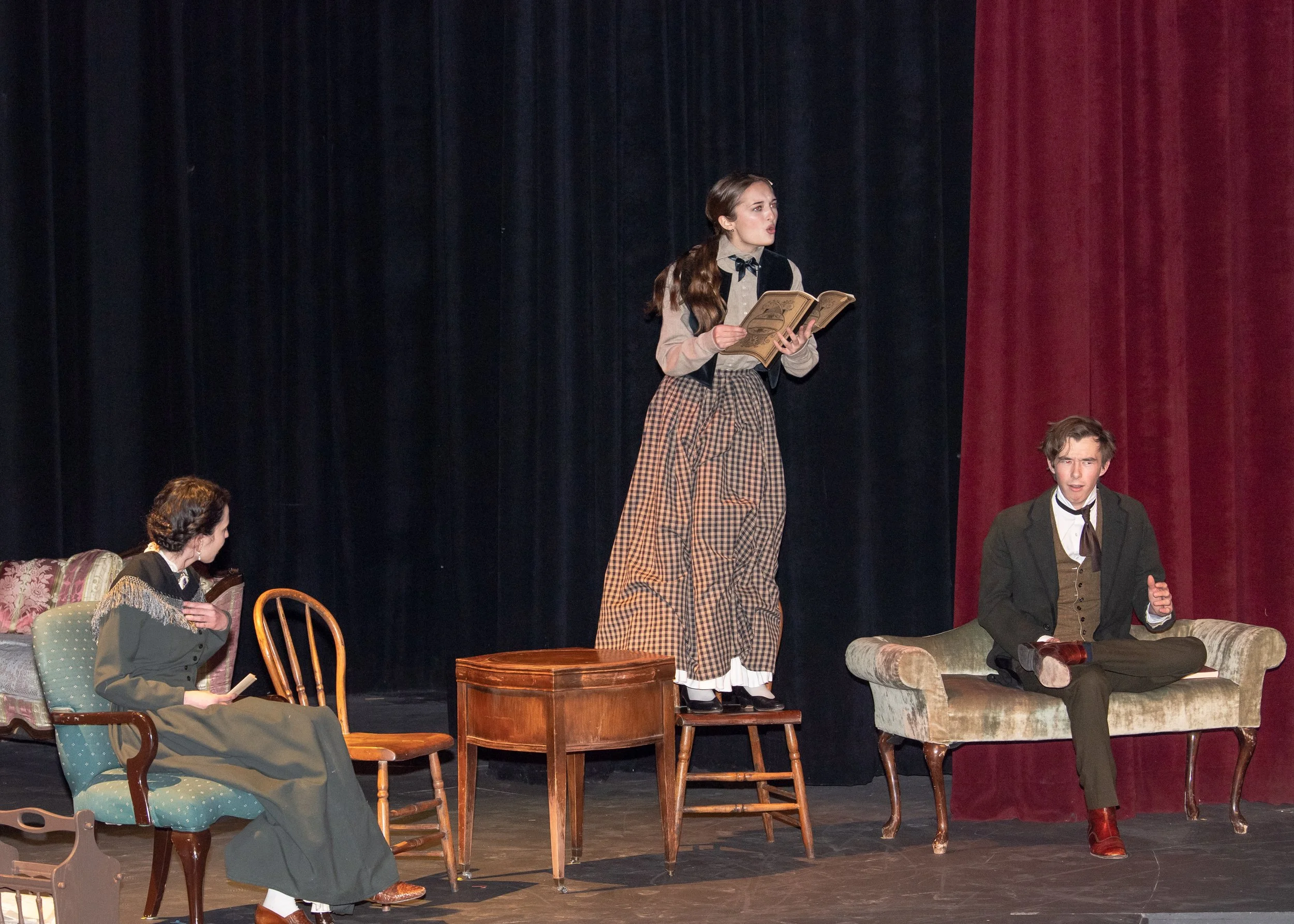 Three young actors on stage in vintage clothing, performing a play. One girl stands on a small stool holding a book, another girl sits on a sofa reading, and a boy sits on a chaise lounge with a book, all around a wooden table with a black curtain backdrop and red stage curtains.