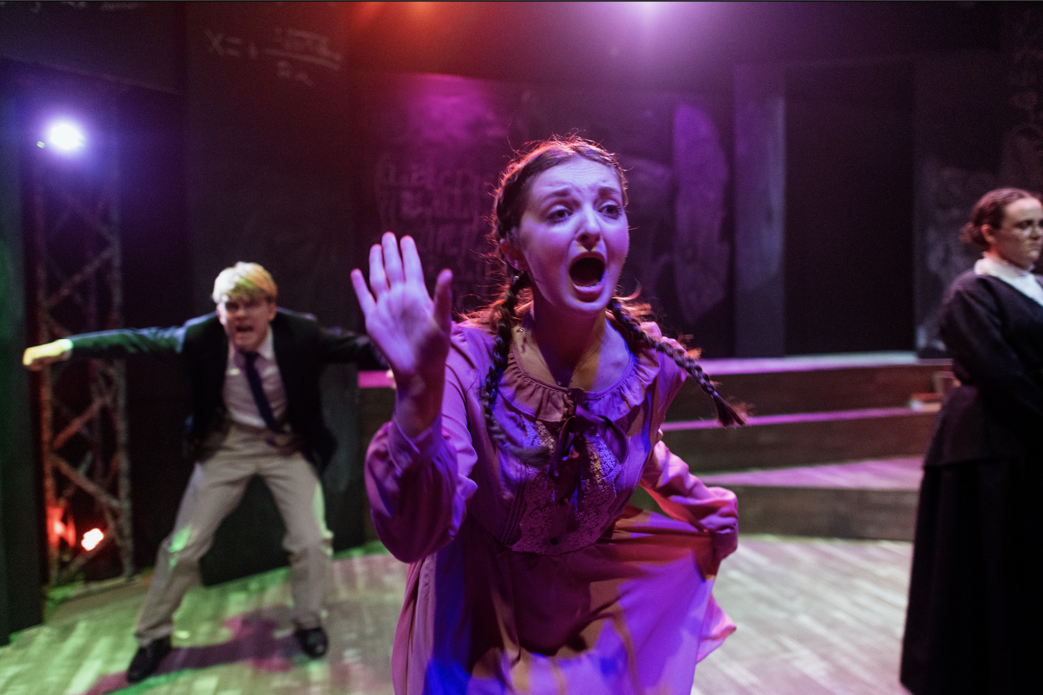A theatrical scene with three actors. The woman in the foreground has braided hair, an expressive face, and is wearing a purple dress. She appears to be shouting or crying out, with her hand raised. Behind her are two other actors; one is a man in a black jacket and white pants, leaning forward with a distressed expression, and the other is a woman in a dark outfit with glasses, standing with a serious look. The background features stage lighting and set pieces.