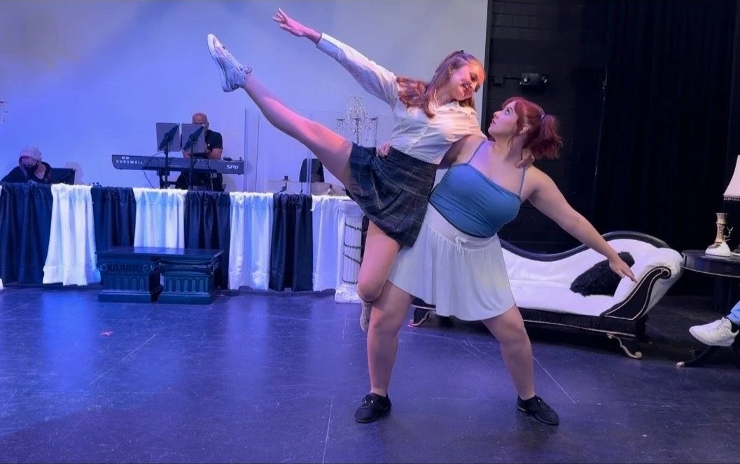 Sarah Novak in Cruel Intentions at Shifted Lens Theatre Company. Two women with one with their leg in the air being lifted by the other. They are performing in a show, in school clothes. In front of a white and black couch on a stage.