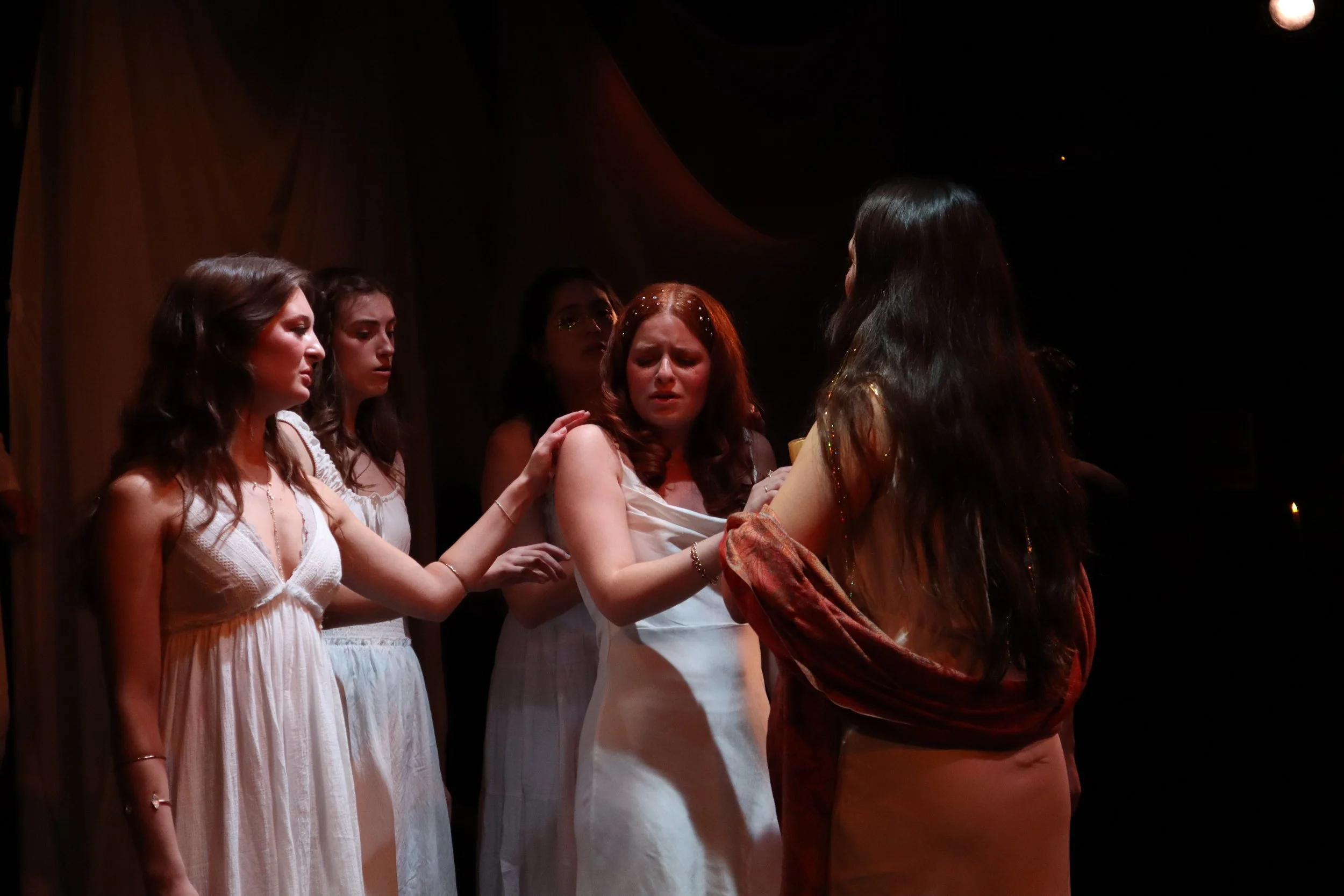 A group of women dressed in white and beige gowns on a stage, with one woman in a dark dress, appears to be performing a dramatic scene.