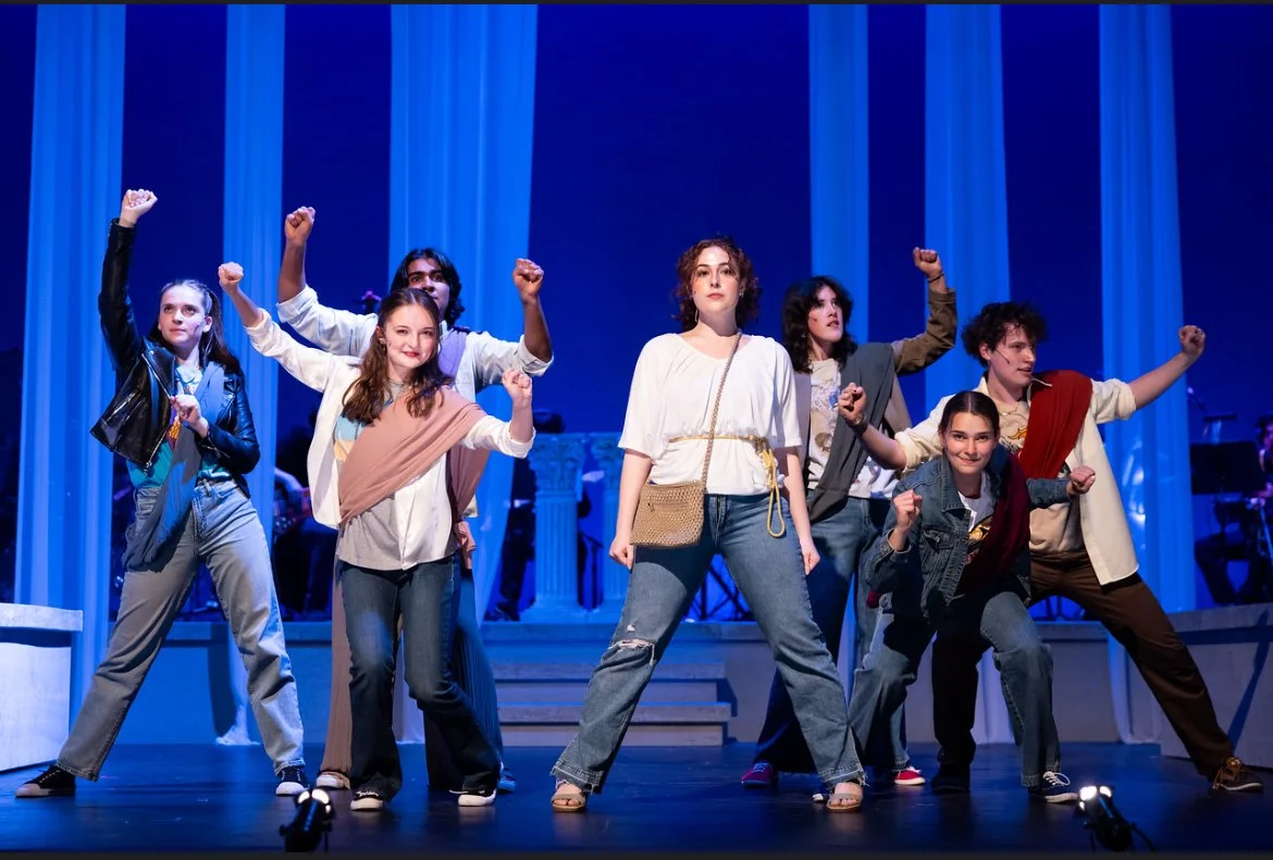 A group of young performers on stage, striking confident poses with fists raised, during a theatrical production with a blue and white backdrop.