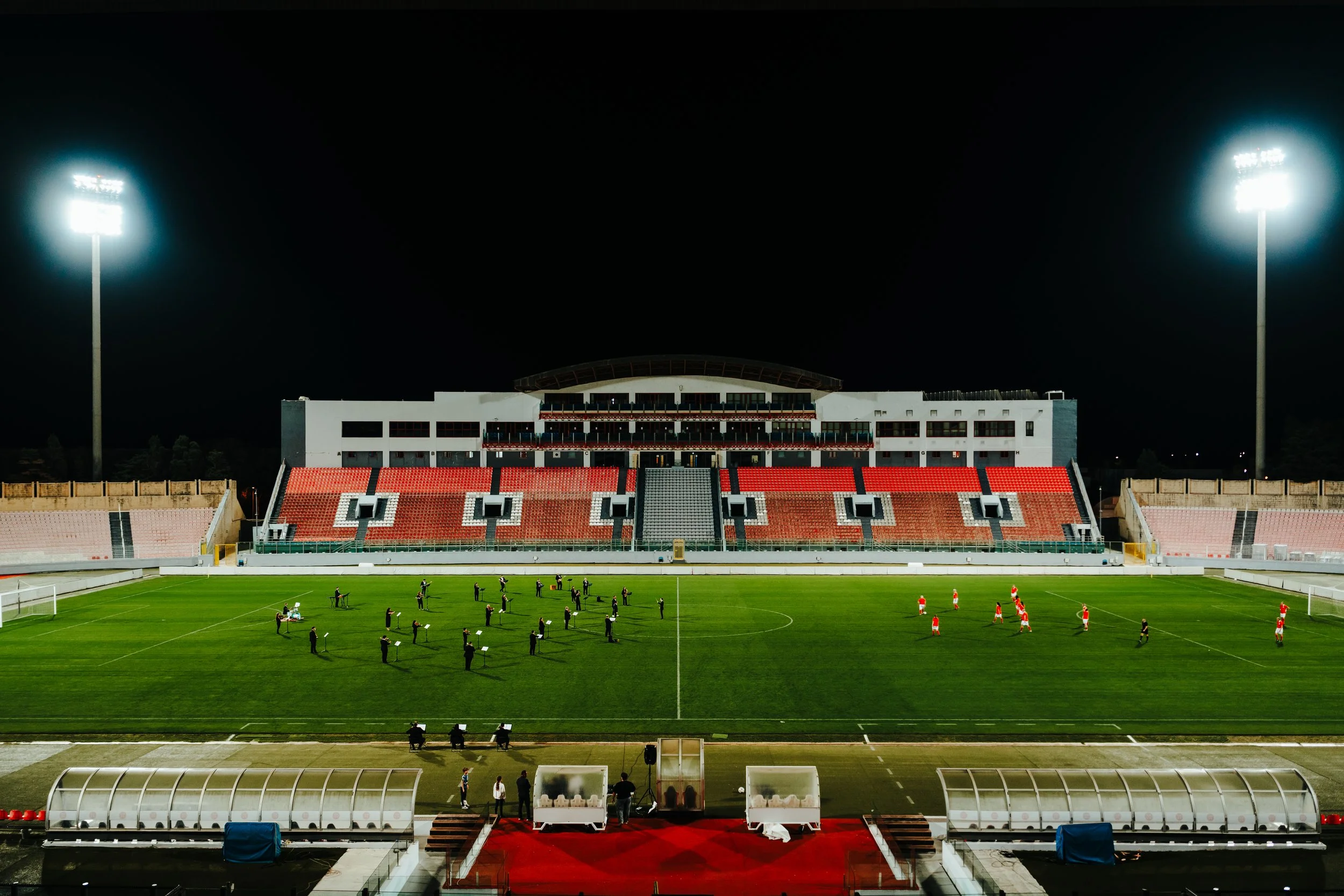 Nighttime view of a soccer stadium with bright floodlights, red and black seating sections, and a group of people standing on the field, featuring a 30 piece Musea Orchestra and the Malta National Football teams performing in the stadium.