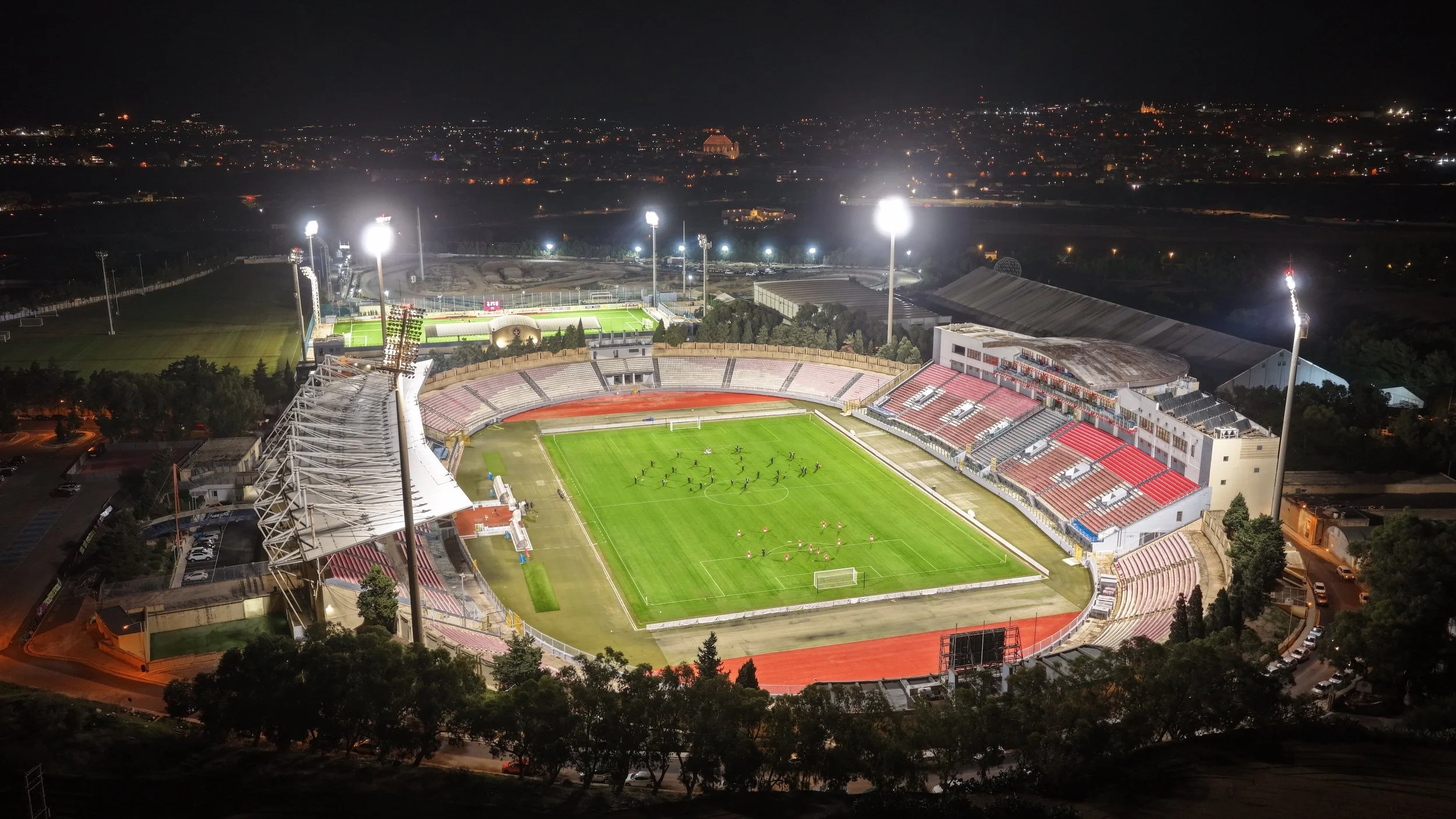 Aerial night view of a brightly lit soccer stadium with green field, red running track, and seating sections, surrounded by trees and city lights featuring a 30 piece Musea Orchestra and the Malta National Football teams performing in the stadium.