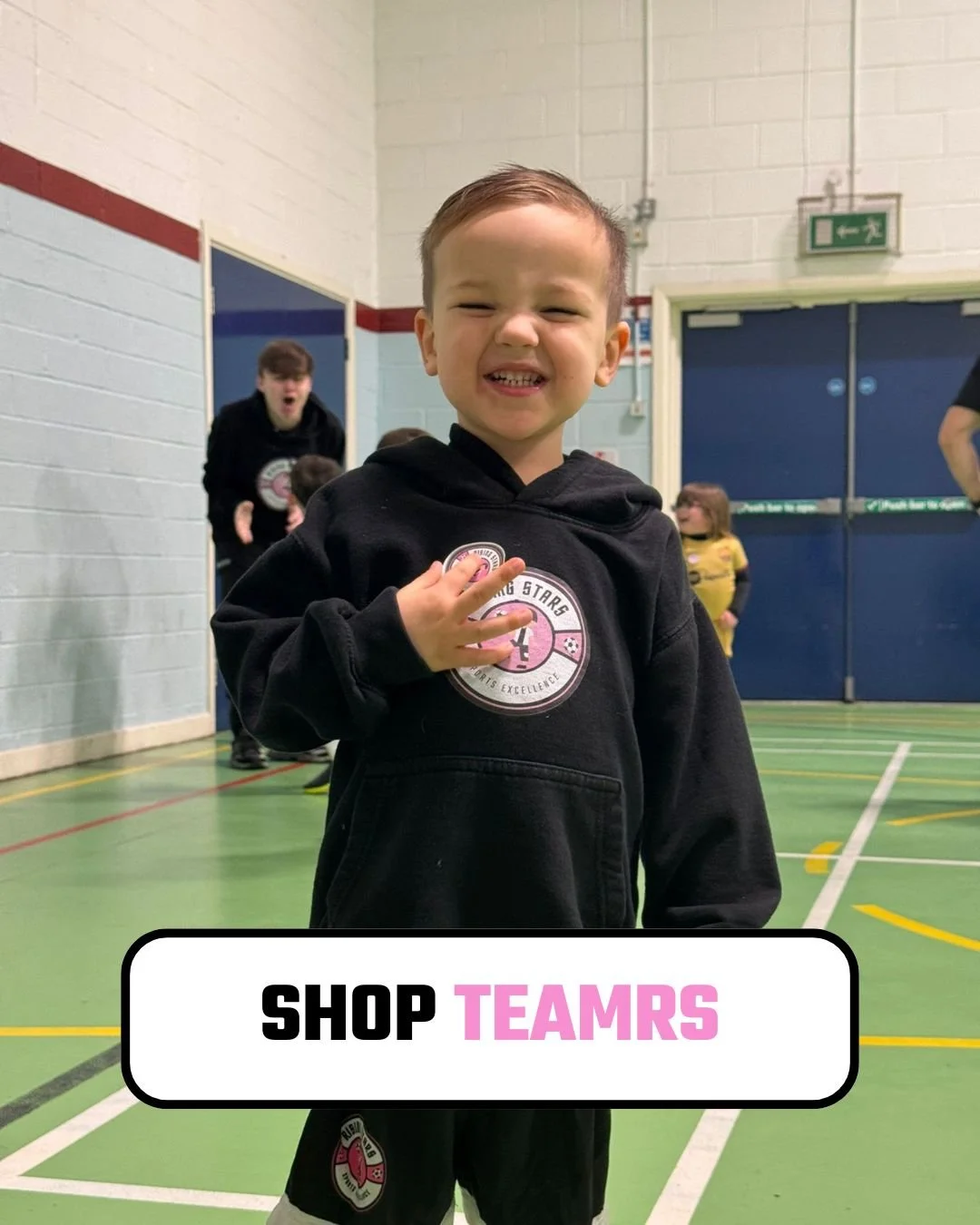 A young boy in a black hoodie with a sports logo, smiling and making a peace sign, standing in a gymnasium with other children and a coach in the background.