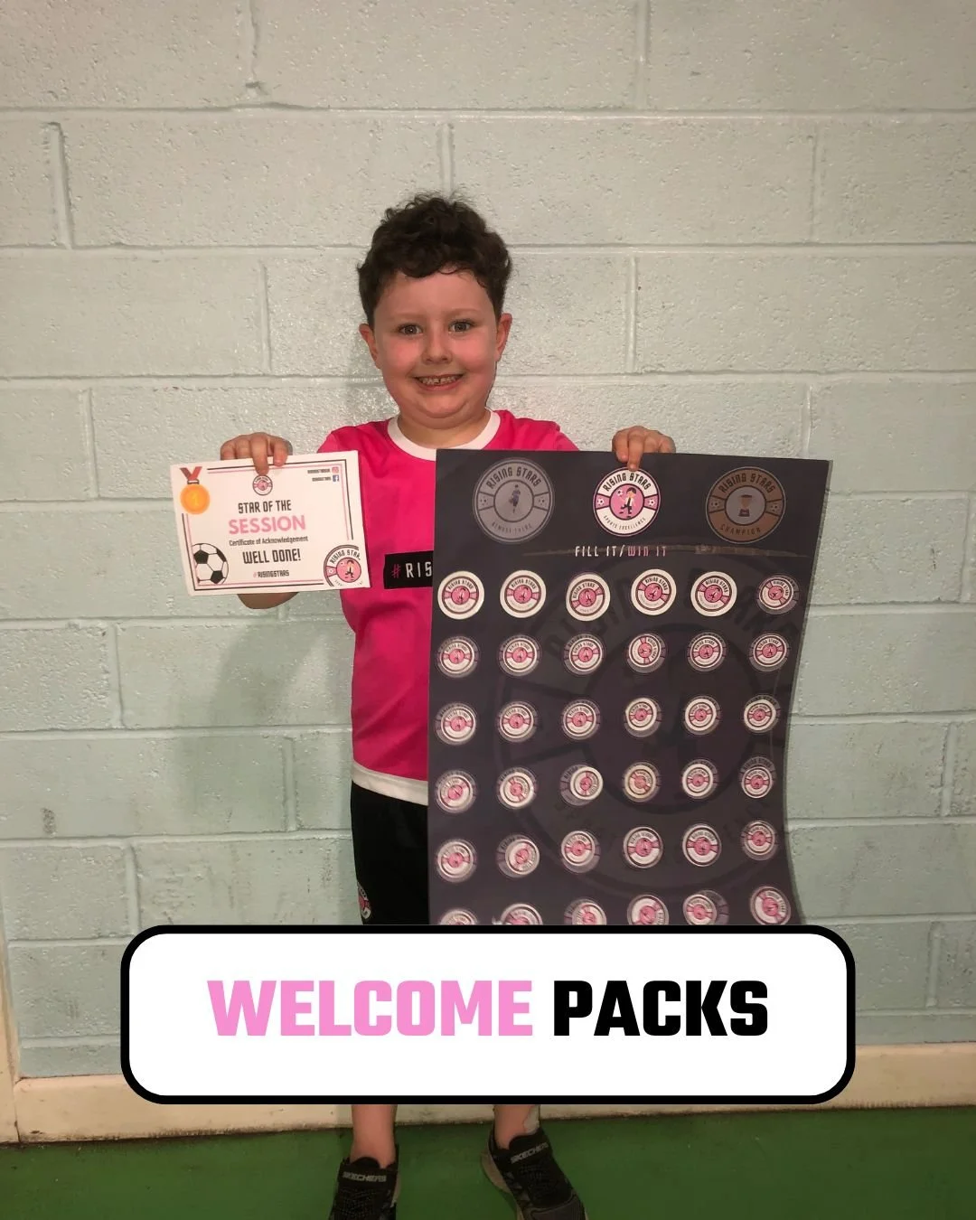 A young boy with short curly hair smiling and holding a small certificate in his right hand and a large poster board with stickers in his left hand, standing against a light gray brick wall.